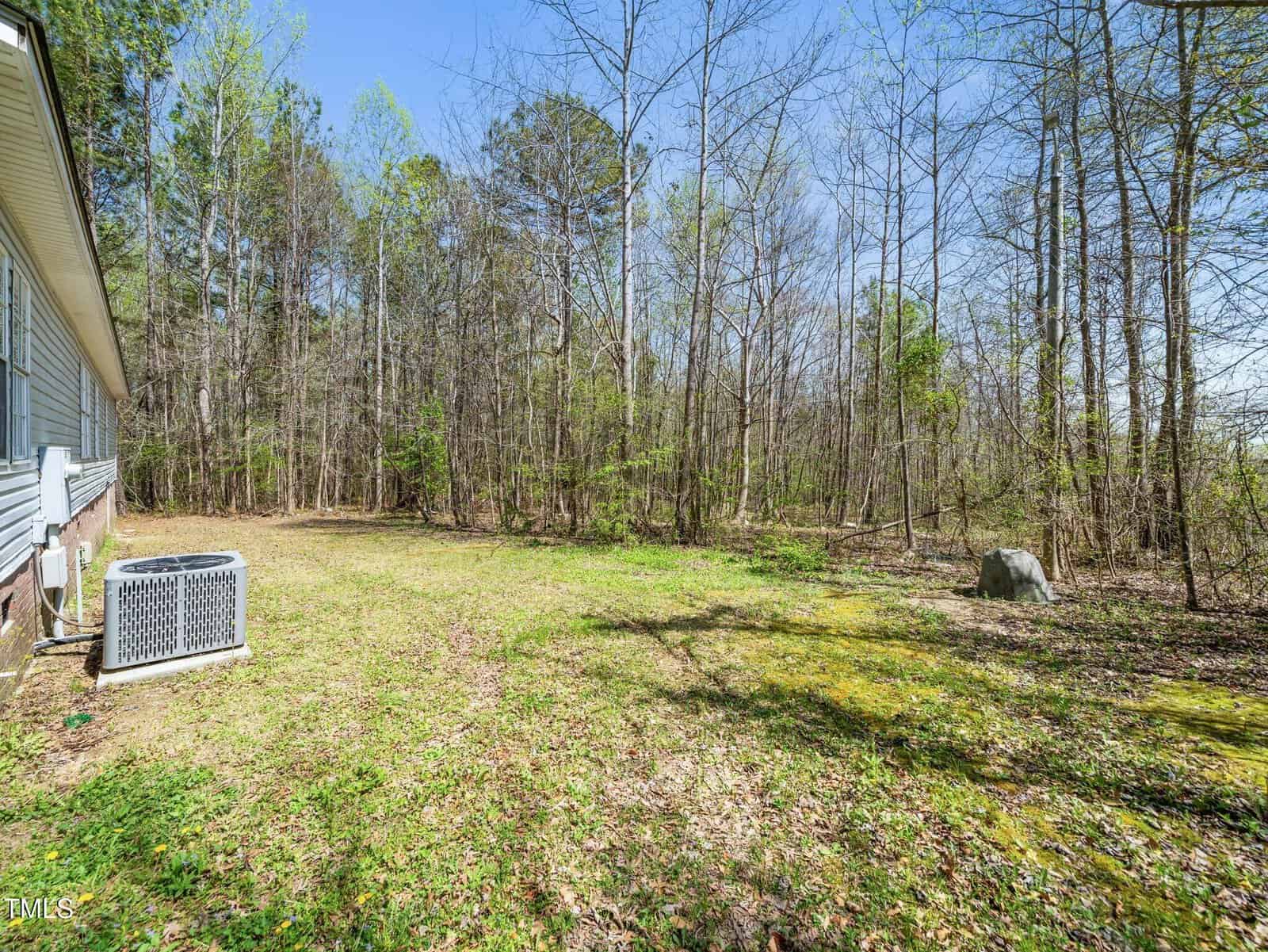 A grassy backyard bordered by woods, with an HVAC unit near the house on the left and a large rock on the right. Trees are sparse with early spring foliage, and sunlight casts shadows on the ground.