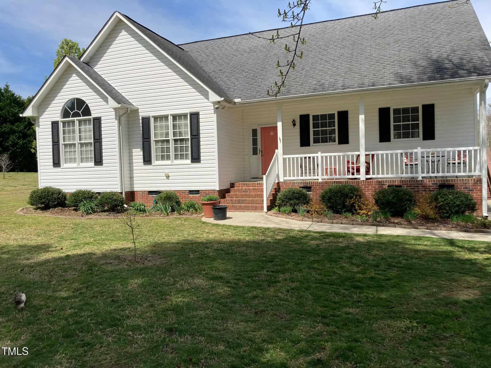 Nestled at 126 W Quail Ridge Road, Oxford, this single-story white house boasts a gray roof and black shutters, while a welcoming front porch features a swing and vibrant red chairs. The manicured lawn and small shrubs guide you along the walkway under a clear blue sky.
