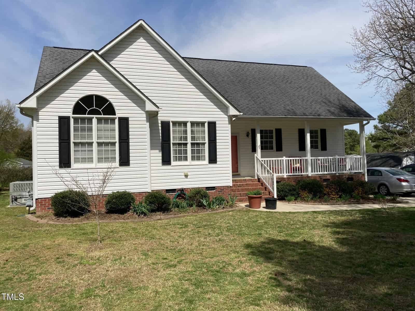 A white single-story house with black shutters, a dark roof, and a covered front porch. The house is surrounded by a grassy yard with shrubs and a small tree. A car is parked in the driveway on the right.