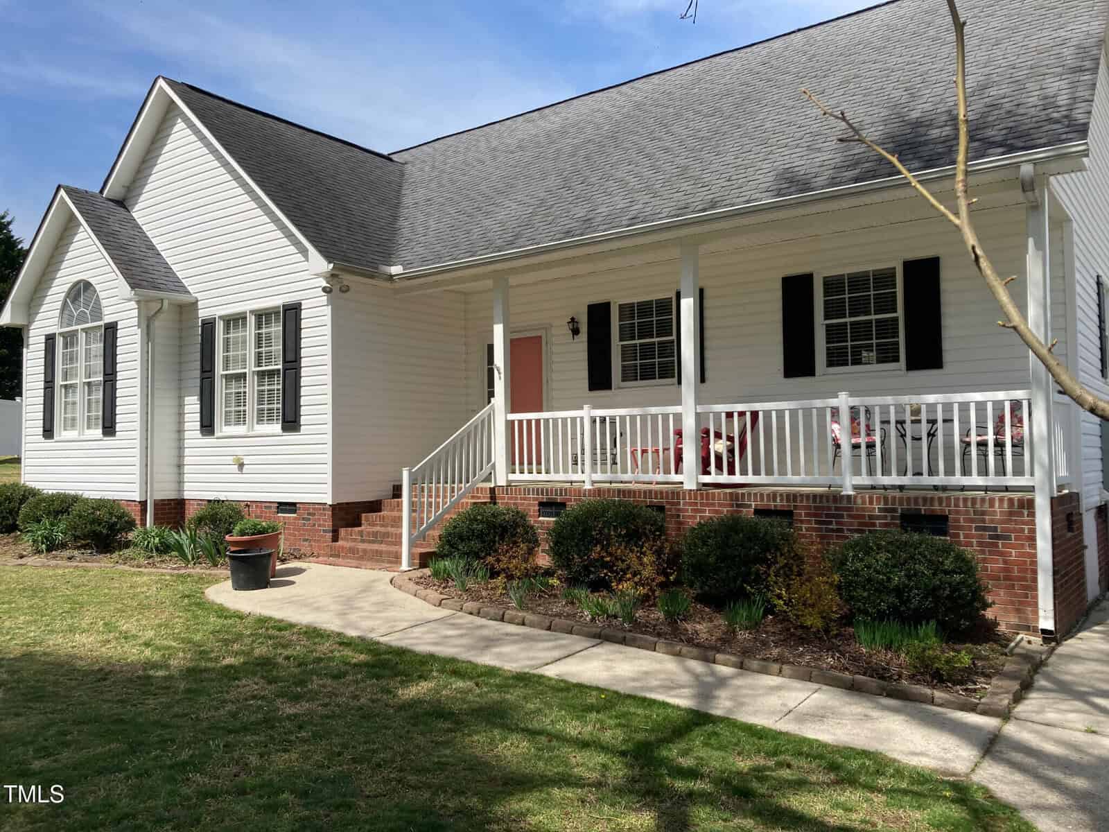 A single-story white house with a gable roof and red brick foundation. It features black shutters, a red front door, and a covered porch with white railing. The yard is landscaped with shrubs and a potted plant near the entrance.