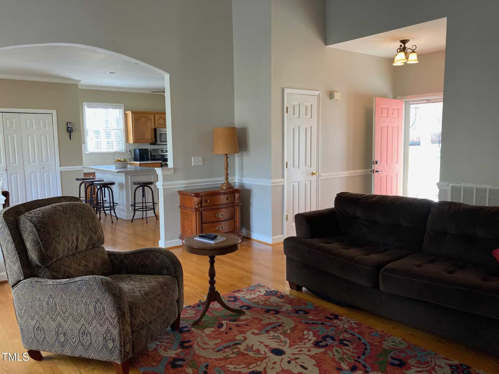 A living room with a brown sofa, patterned armchair, wooden coffee table, and colorful rug. An open doorway leads to a kitchen with a bar counter, stools, and white cabinets. A pink door is slightly open, allowing sunlight to enter the room.