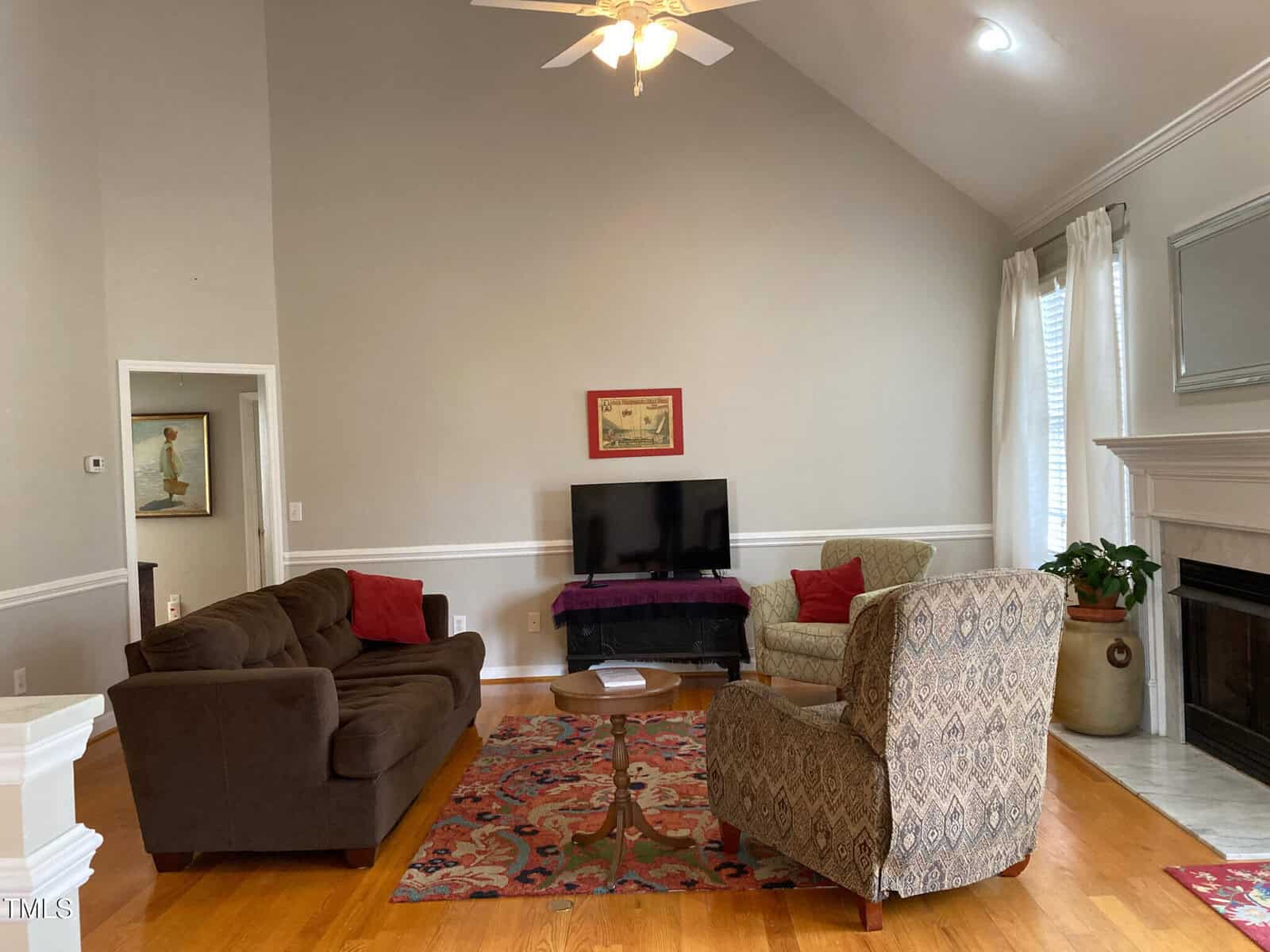 A living room with a high ceiling features a couch, two patterned chairs, a TV on a stand, and a colorful rug. There are potted plants, a fireplace, and a ceiling fan. The walls are light gray and a window with white curtains is on the right.