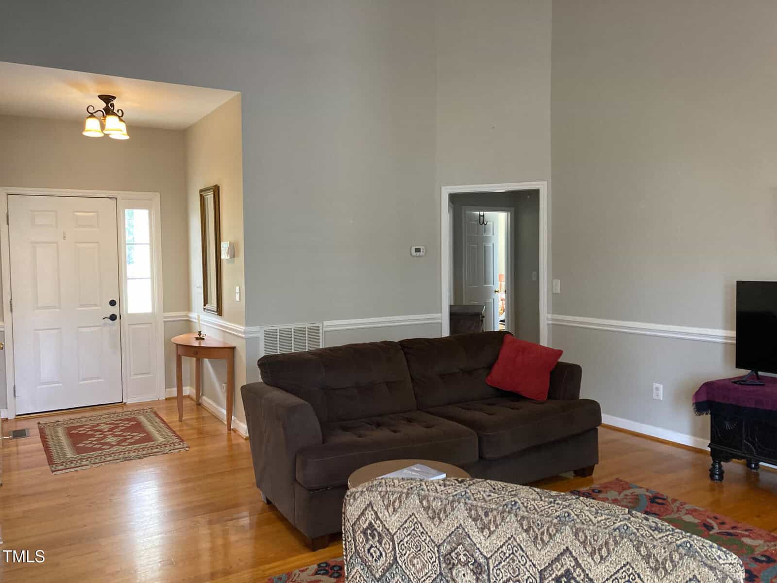 Living room with a brown sofa, red and white cushions, and a patterned ottoman. A small TV is on a stand to the right. There is a console table near the white front door. Hardwood floors and a light fixture on the ceiling are visible.