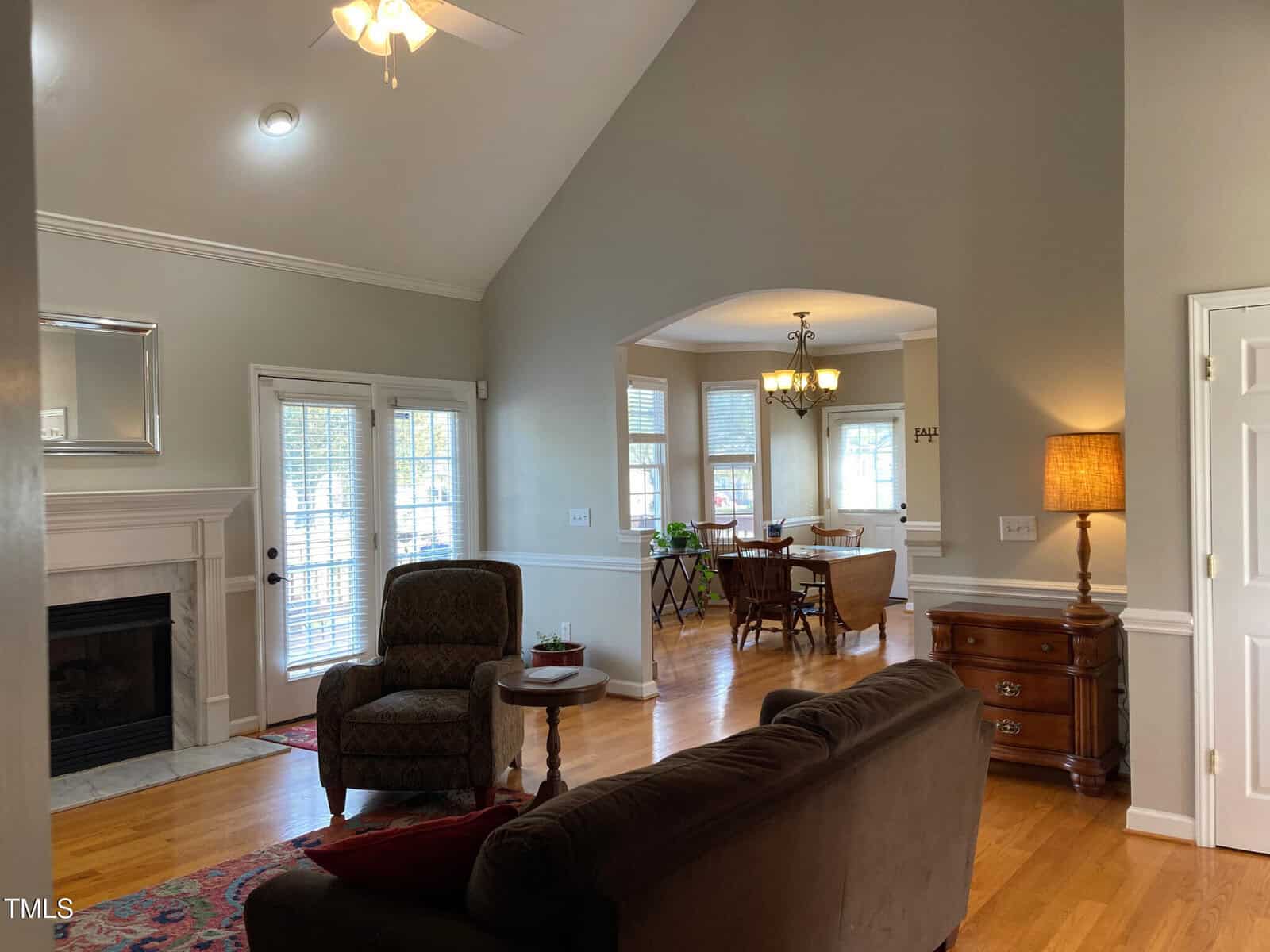 A living room with hardwood floors and neutral walls, featuring a couch, an armchair, a fireplace, and a ceiling fan. In the background, a dining area with a round table and chairs is visible. A table lamp is on a wooden side table.