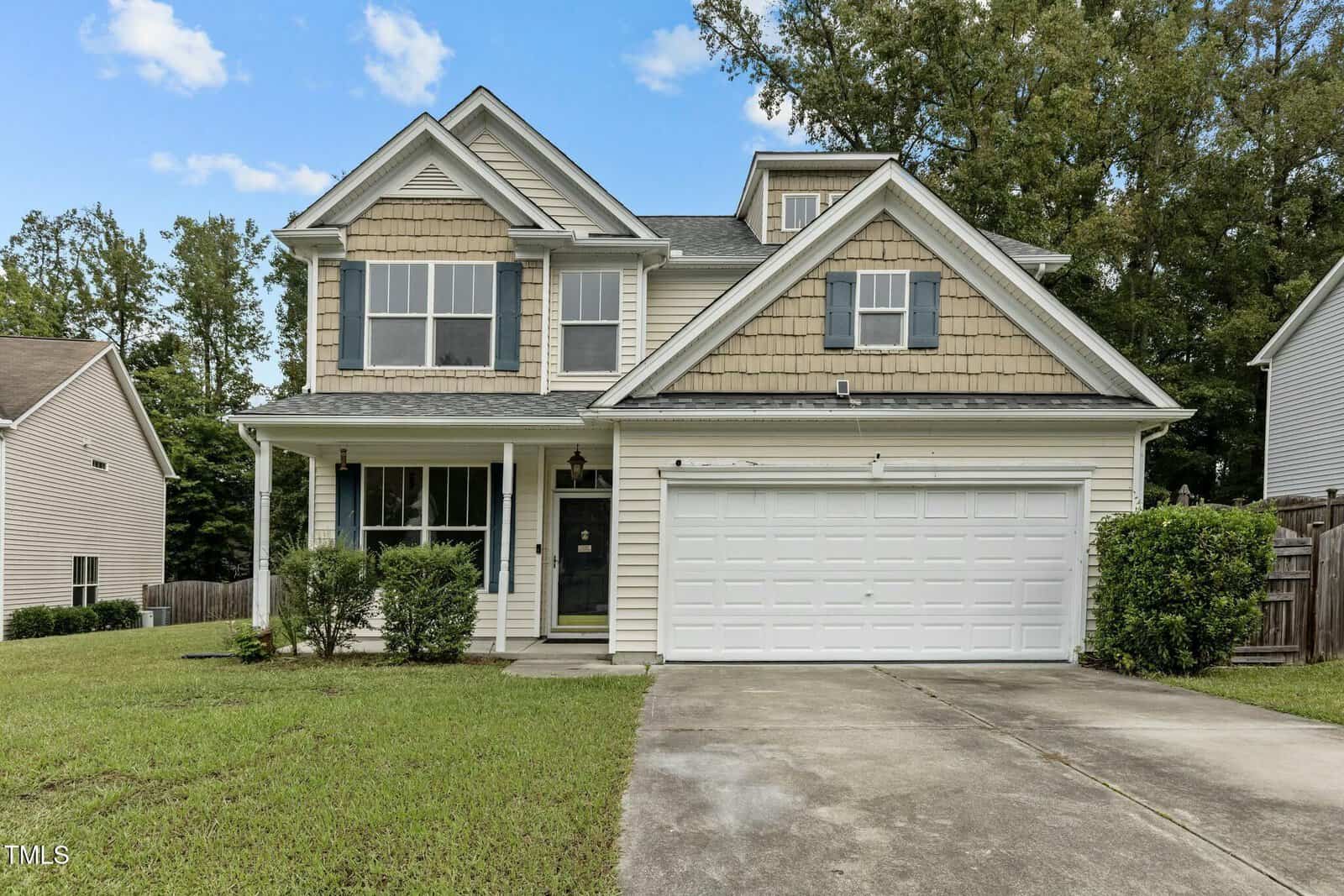 Located at 2112 Queensway Court, this two-story suburban house in Creedmoor features beige siding and a gray roof. The front boasts a double garage, a cozy porch, and manicured bushes. A concrete driveway completes the scene under clear skies with scattered clouds.
