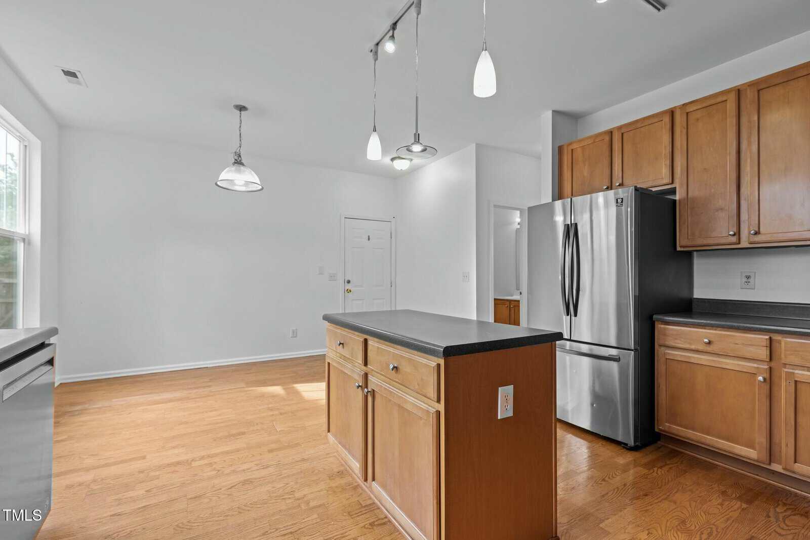A spacious kitchen with wooden cabinets, a central island, and stainless steel appliances. The room has hardwood floors and overhead pendant lighting. There is a door leading outside and natural light from a large window.