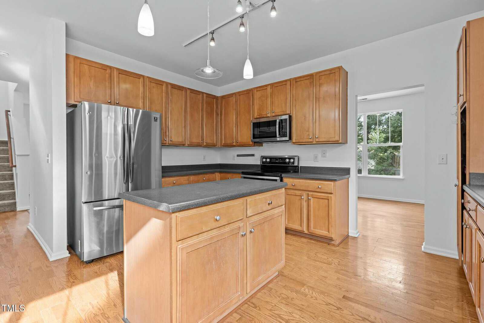 A modern kitchen with wooden cabinets, stainless steel appliances, and a central island. The floor is hardwood, and there are pendant lights above the island. A large window provides natural light from the right side.