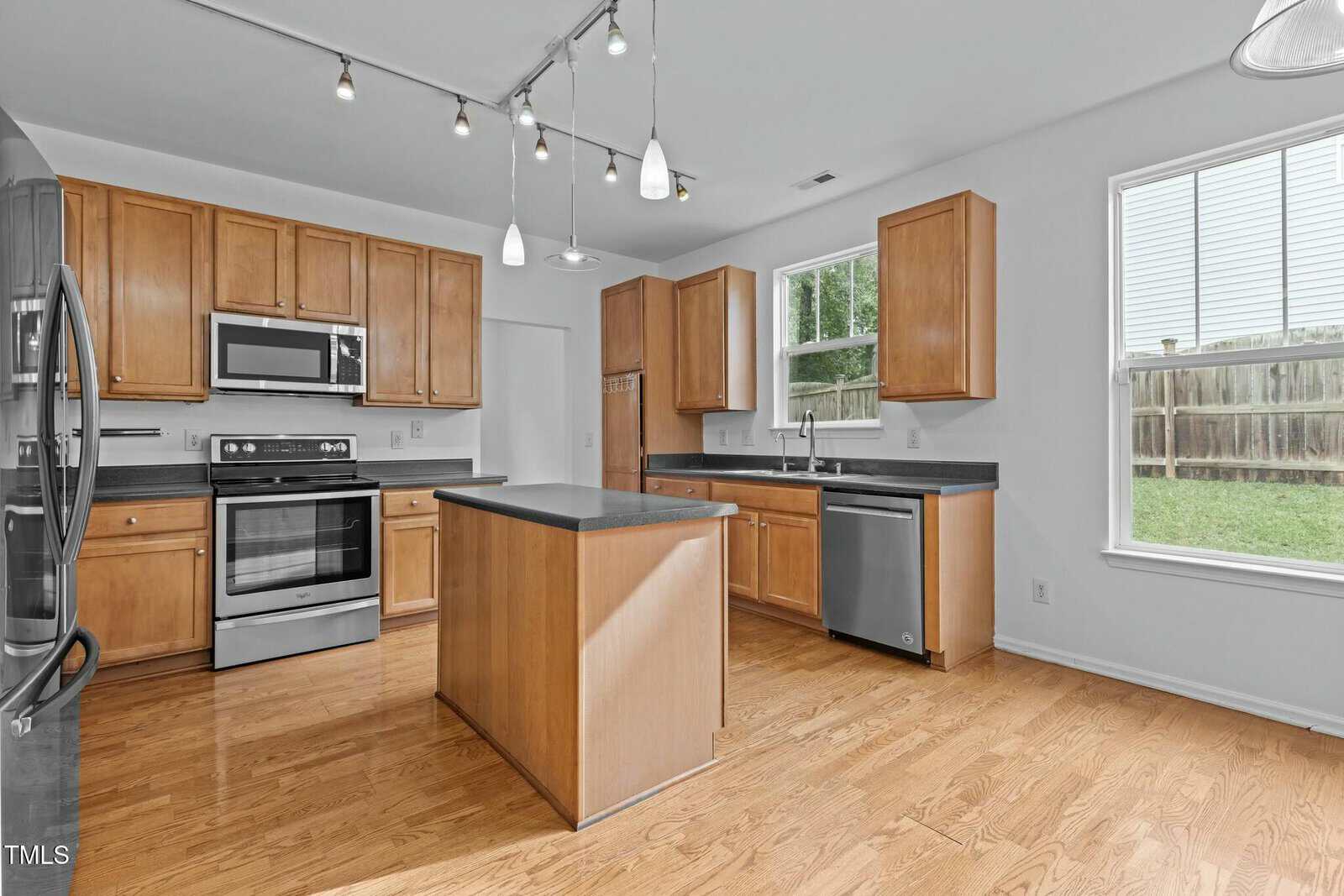Modern kitchen with wood cabinets, black countertops, and stainless steel appliances, including a refrigerator, oven, microwave, and dishwasher. An island is centered under pendant lighting, and a window offers natural light. Hardwood floors throughout.