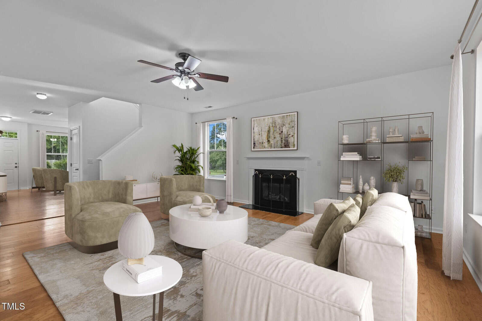 A modern living room with a beige sofa, two olive-green chairs, and a round coffee table. A fireplace is centered against the wall, with artwork above. There's a ceiling fan, large windows with curtains, and a plant in the corner.