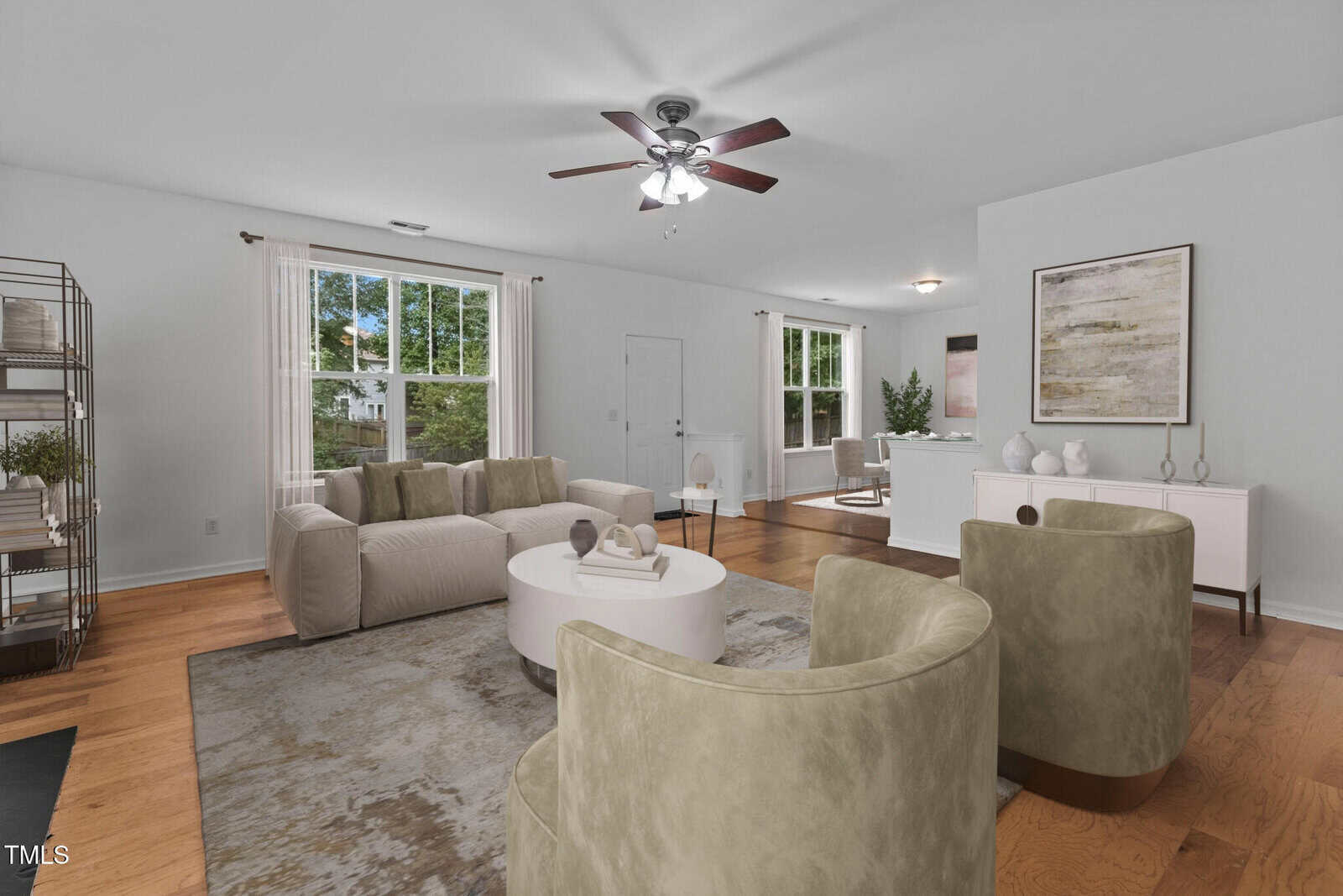 A spacious living room with light-colored walls and wooden flooring. It features a beige sofa, two green armchairs, a round white coffee table, and a ceiling fan with lights. Large windows allow natural light to fill the room.