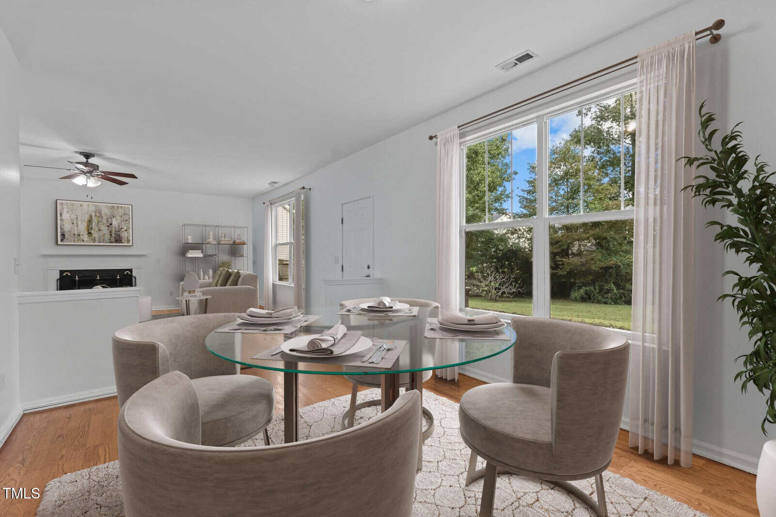 A dining area features a round glass table set for four with gray upholstered chairs on a light-patterned rug. Large windows with sheer curtains reveal a view of a grassy outdoor scene. A glimpse of a living room with a ceiling fan is visible.