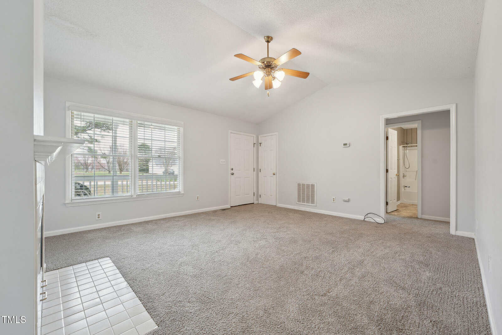 Empty living room with beige carpet, white walls, a ceiling fan with lights, a tiled fireplace hearth, large window, double doors, and an open doorway leading to a bathroom. The room has a vaulted ceiling.