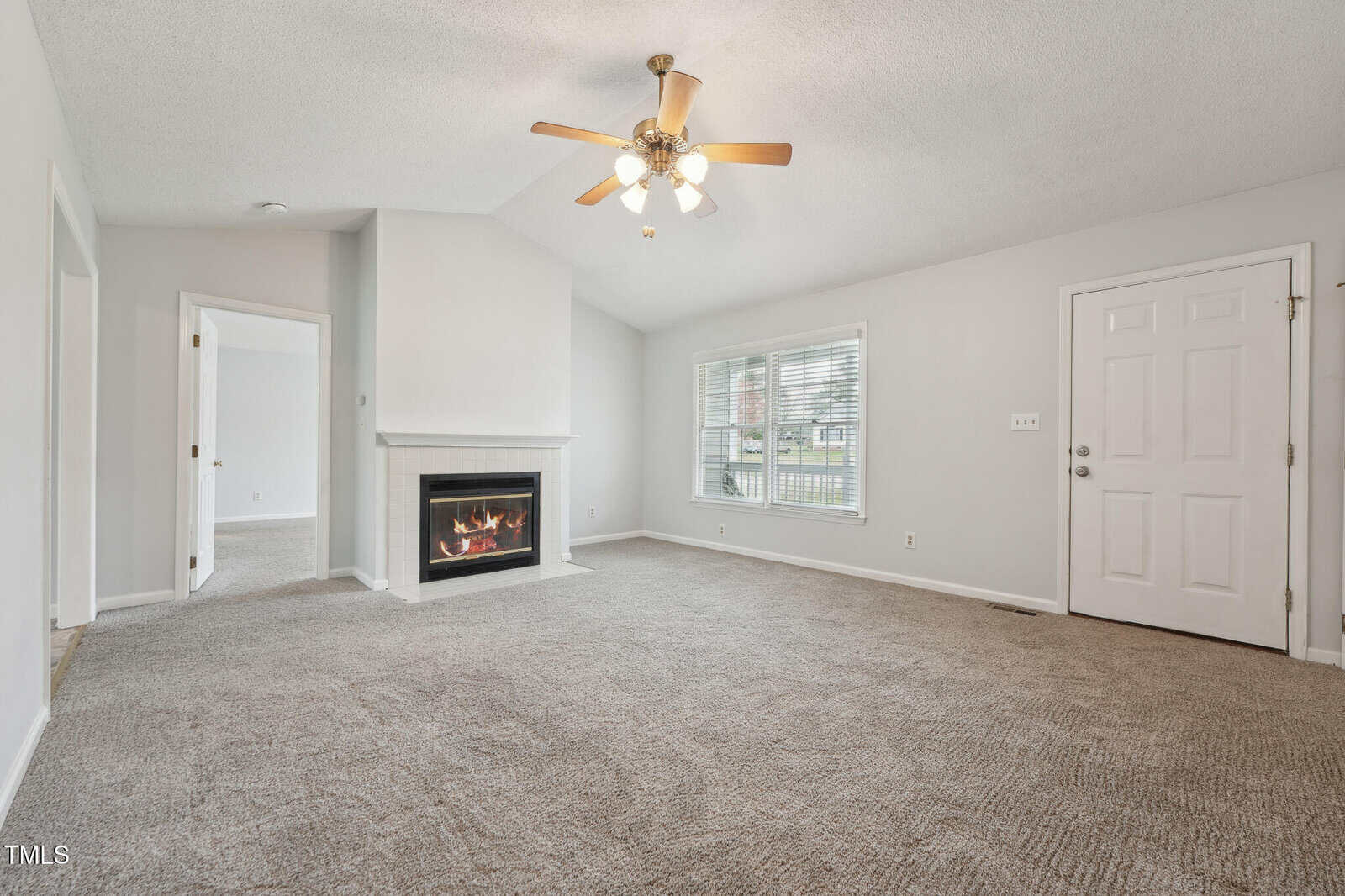 A carpeted living room with light gray walls, a white front door, a window with blinds, a ceiling fan with lights, and a corner fireplace with a lit fire. An open doorway and room are visible to the left.