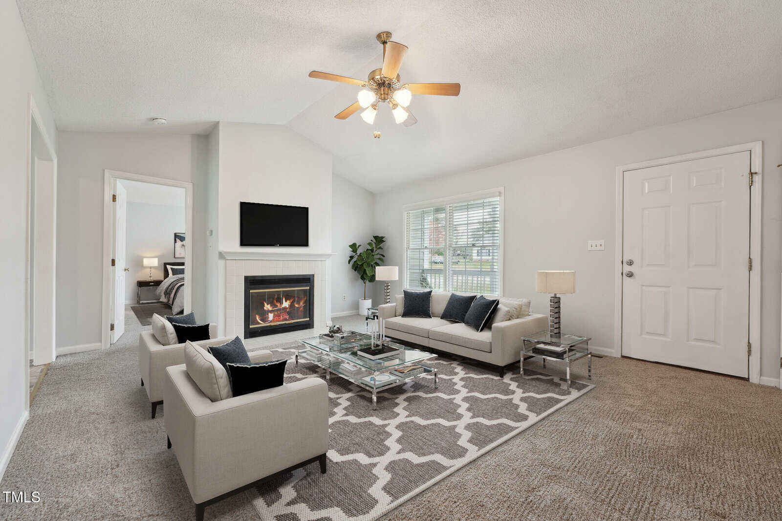 A living room with light-colored walls, two beige sofas, a glass coffee table, a gray patterned rug, a fireplace with a TV above it, and a ceiling fan; windows let in natural light.
