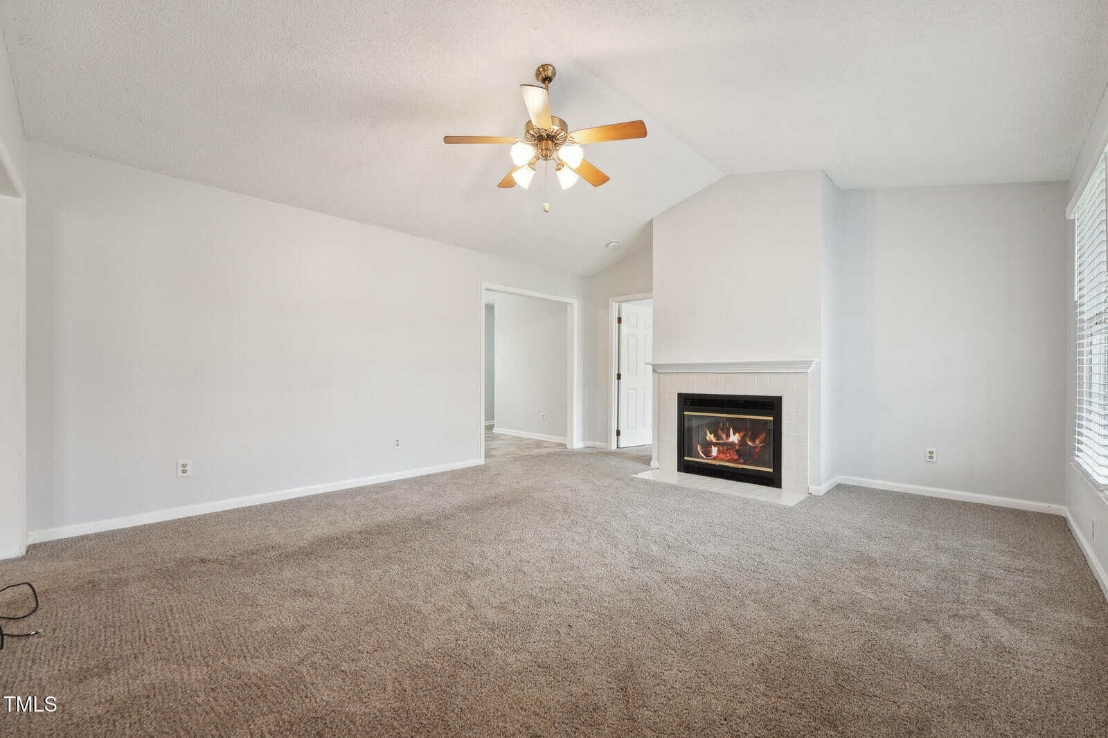 A spacious living room with beige carpet, white walls, a ceiling fan with lights, a corner fireplace, and large windows letting in natural light. An open doorway leads to another room in the background.