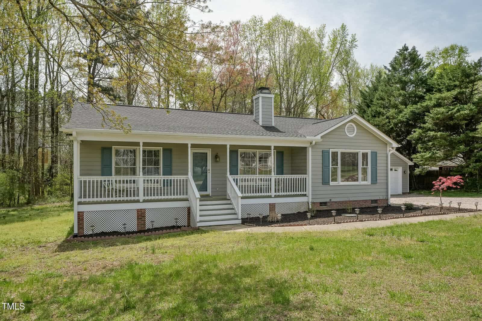 Single-story house with light blue siding, white trim, a front porch with railings, and steps leading to the entrance. The yard has grass and some landscaped beds, surrounded by trees and a driveway on the right side.