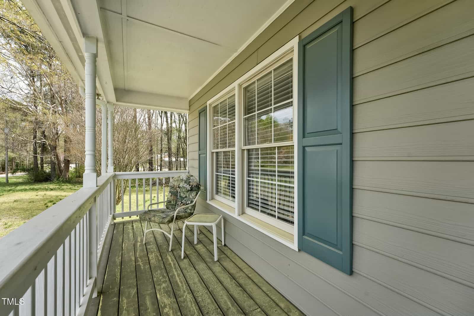 A wooden front porch with light gray siding and blue shutters, featuring two chairs and a small table. A white railing borders the porch, and trees are visible in the yard beyond.