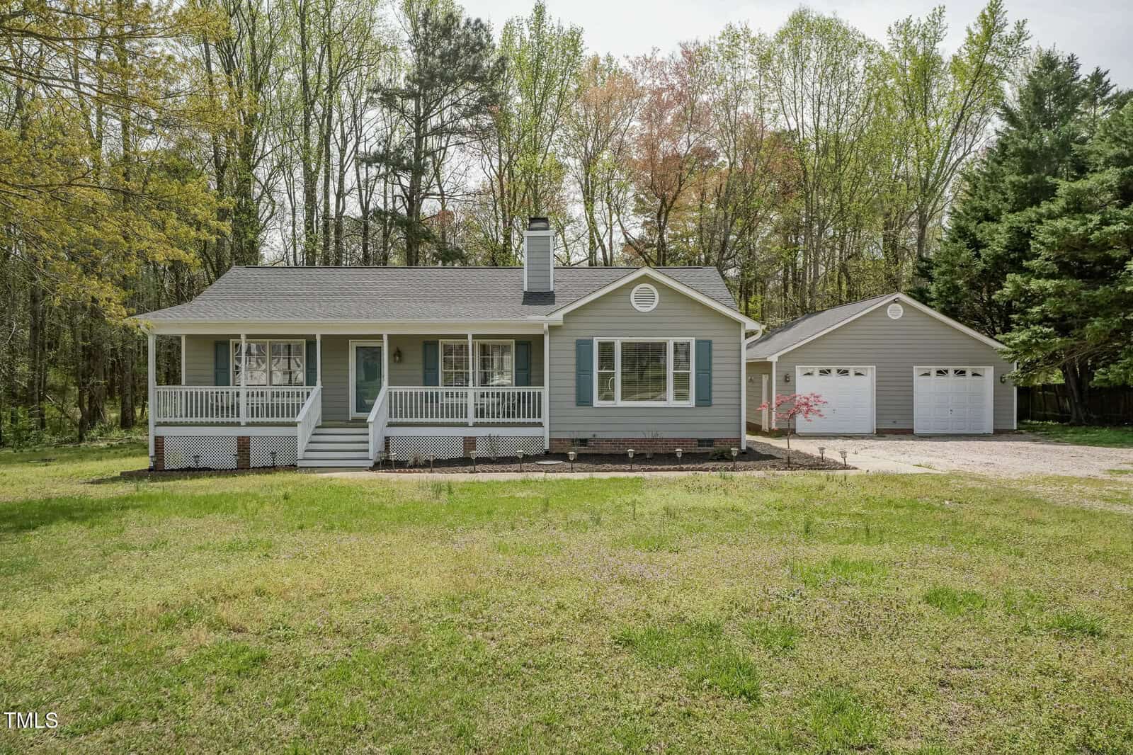 Single-story gray house with white trim and front porch, surrounded by grass and trees, with a detached two-car garage on the right side under a partly cloudy sky.