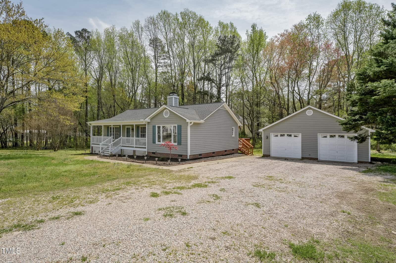 Single-story gray house with a covered front porch and detached two-car garage, surrounded by trees. The house and garage have white trim, and a gravel driveway leads up to them. The yard is mostly grass with some landscaping.