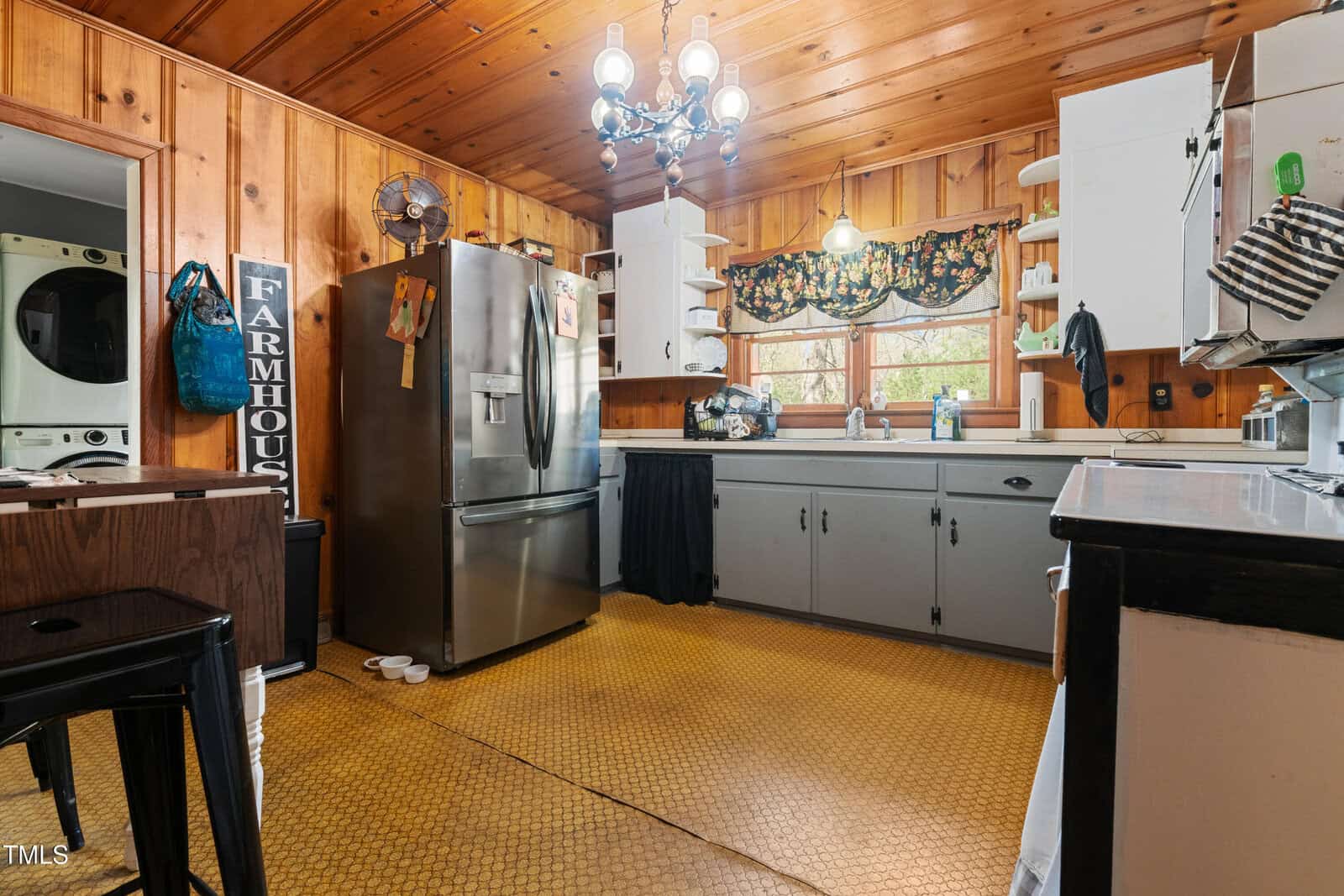 Wood-paneled kitchen with stainless steel fridge, white lower cabinets, open shelves, yellow patterned floor, stacked washer and dryer, farmhouse sign, window above sink, chandelier lighting, and dark curtain under counter.