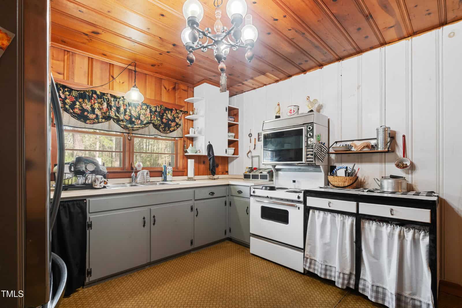 A vintage kitchen with wood panel ceilings, white walls, gray cabinets, a double oven, and open shelving. There is a window with a floral valance, a chandelier, and various kitchen items on the counters and shelves.
