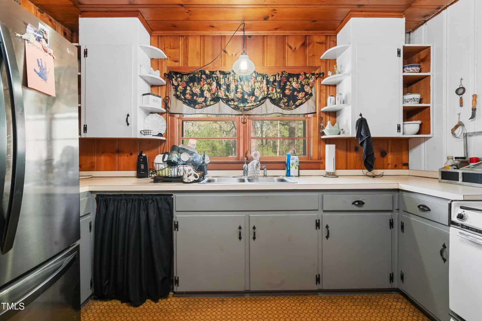 Kitchen with wood-paneled walls, white cabinets, and a double sink under a window. Dishes and cleaning supplies are by the sink. Black skirt covers space under counter. Refrigerator and stove are partly visible.