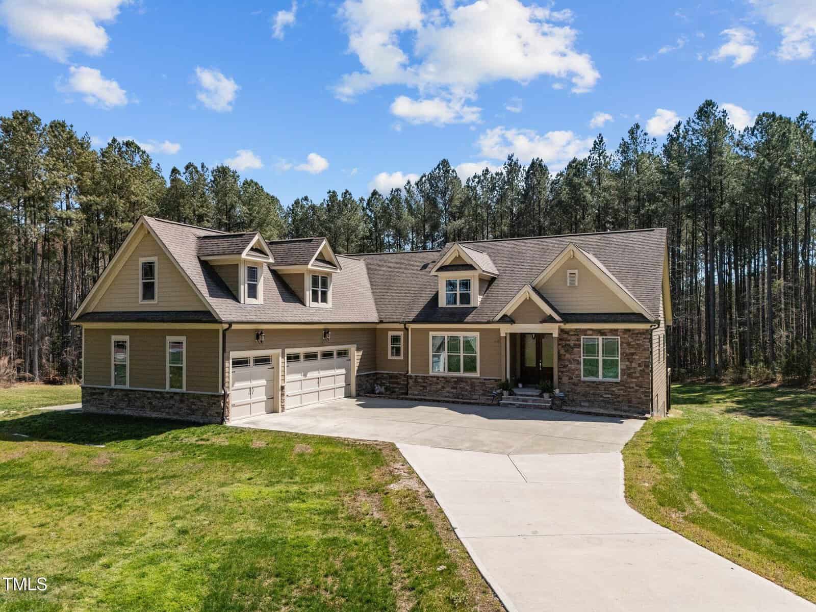 A spacious two-story house at 3220 Brassfield Road in Creedmoor boasts beige siding and elegant stone accents. With three garage doors and a concrete driveway, it's surrounded by a lush lawn and dense forest under a partly cloudy sky.