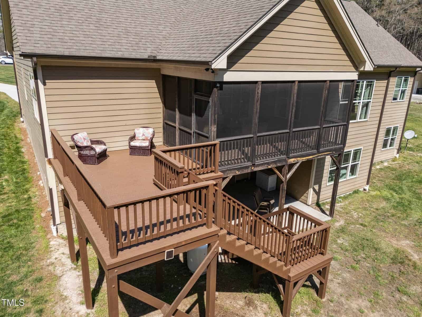 A two-story house with brown siding features an elevated wooden deck with two cushioned chairs. The deck has stair access and a screened porch below. The surrounding area is grassy with a satellite dish visible.