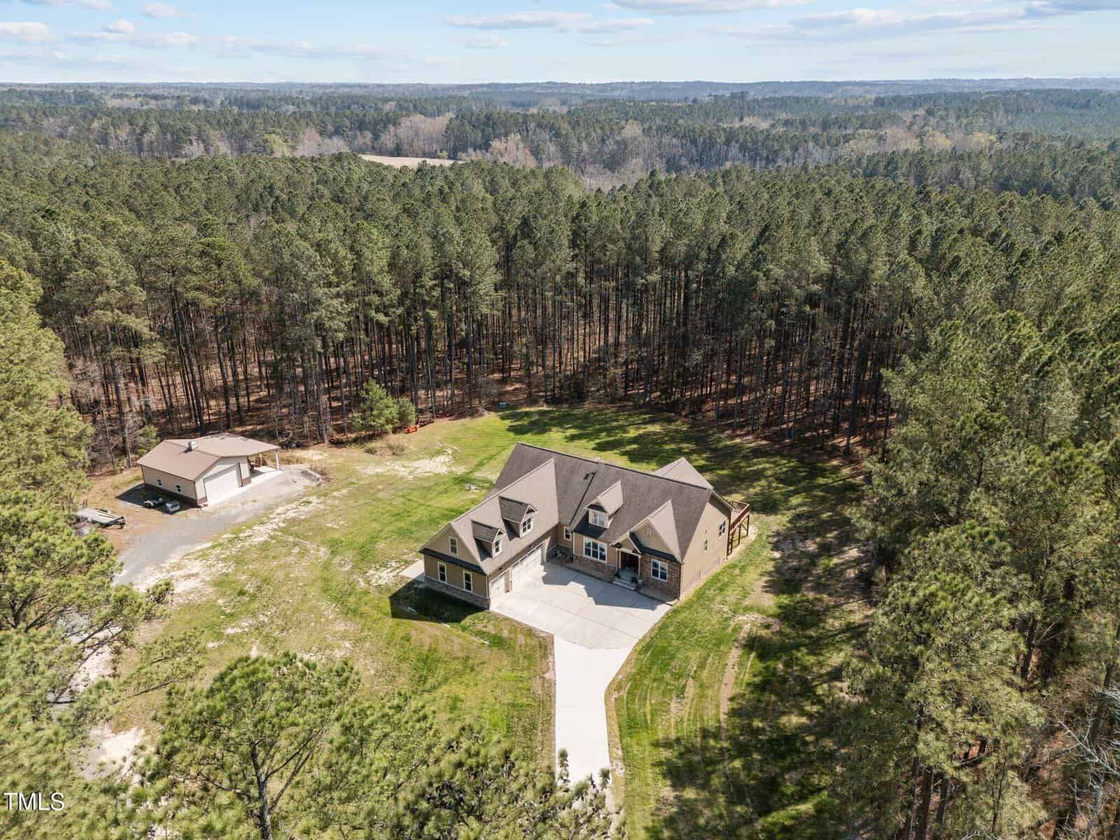 Aerial view of a large house surrounded by dense forest. The home has a gray roof, multiple windows, and a circular driveway. A smaller building is nearby. The landscape features trees, grass, and a clear sky in the background.