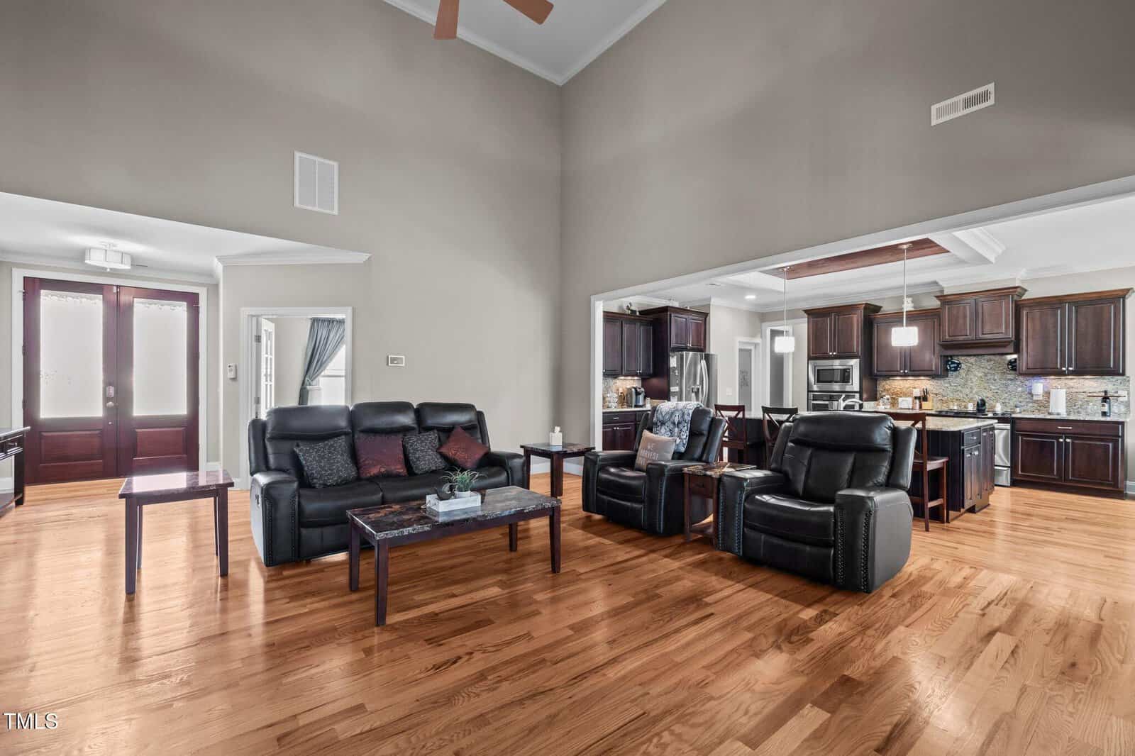 A spacious living room with hardwood floors, black leather seating, a glass coffee table, and side tables, connected to an open kitchen with dark wood cabinets and stainless steel appliances.