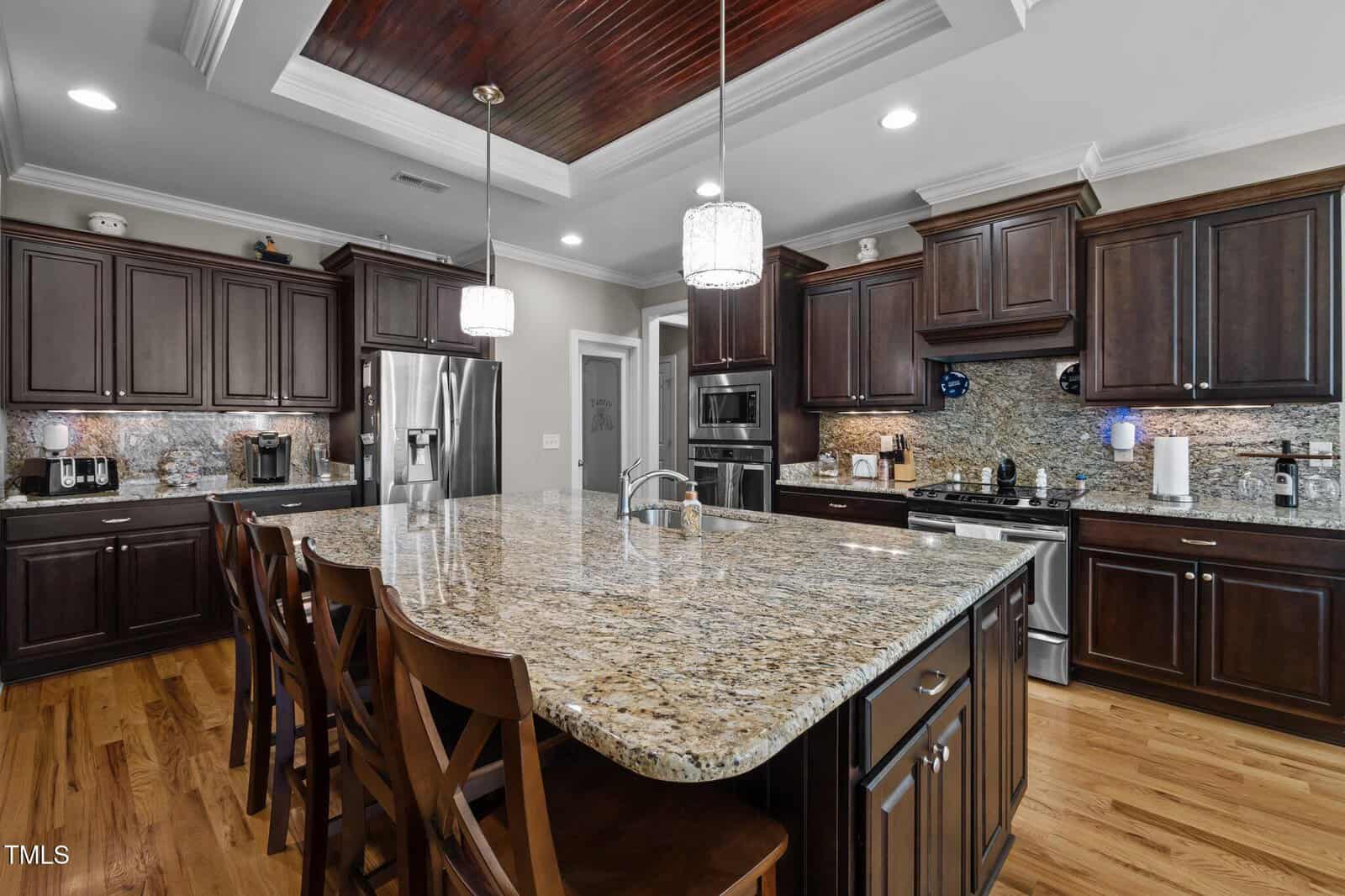 A modern kitchen with dark wood cabinets, stainless steel appliances, granite countertops, a large island with a sink, and four wooden chairs. Pendant lights hang above the island, and the floor is hardwood.