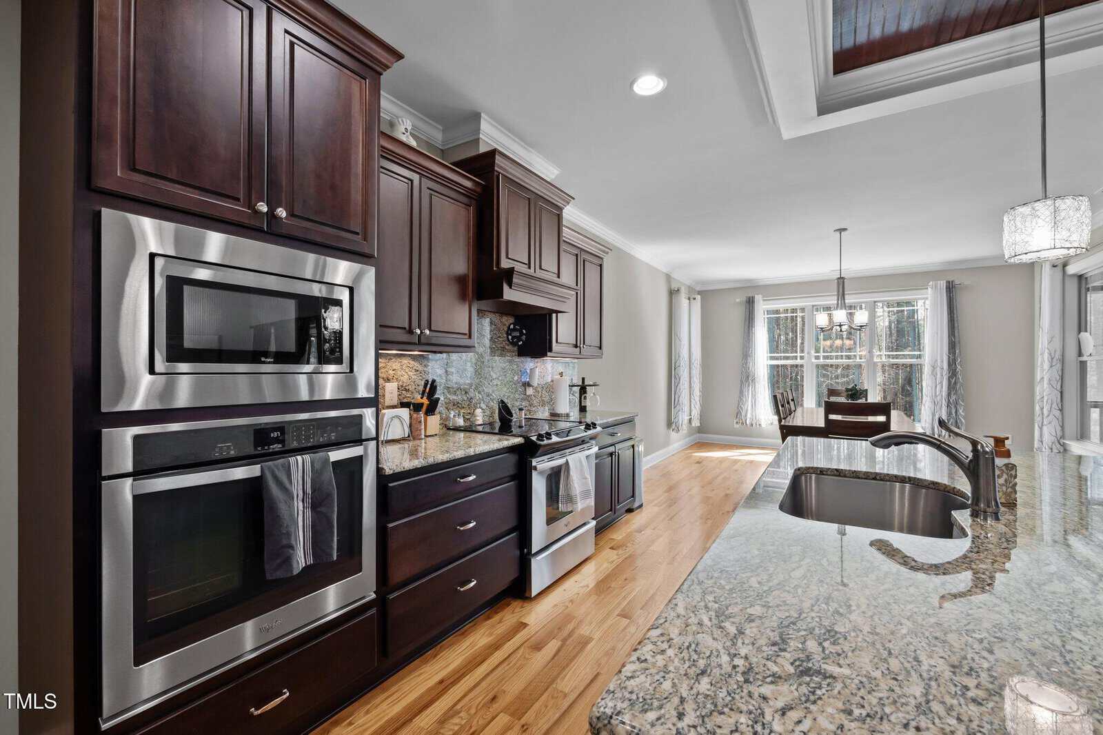 A modern kitchen with dark wood cabinets, stainless steel appliances, a double oven, electric stove, granite countertops, a center island with sink, and hardwood floors. Large windows and a dining area are visible in the background.