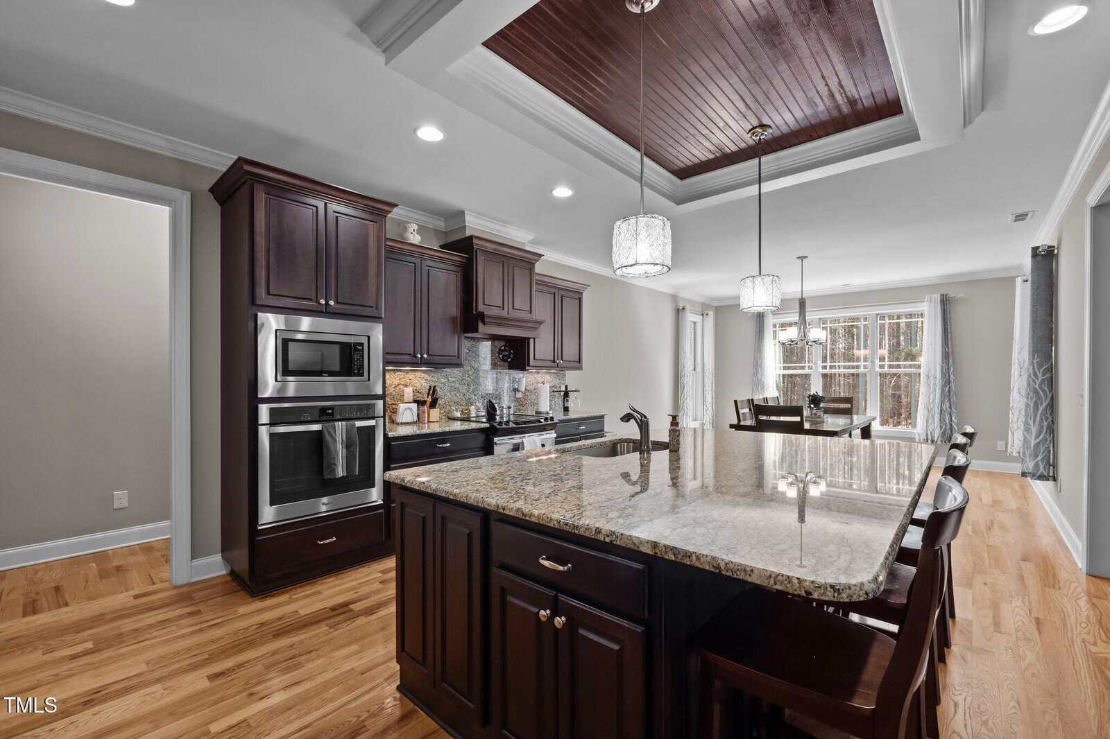 A modern kitchen with dark wood cabinets, stainless steel appliances, a granite island with bar stools, pendant lighting, hardwood floors, and a dining area near large windows in the background.