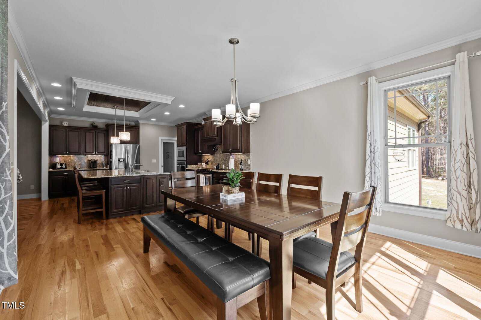 A modern open-concept kitchen and dining area with hardwood floors, dark wood cabinets, stainless steel appliances, a large island, and a dining table with chairs and a bench next to large windows with natural light.