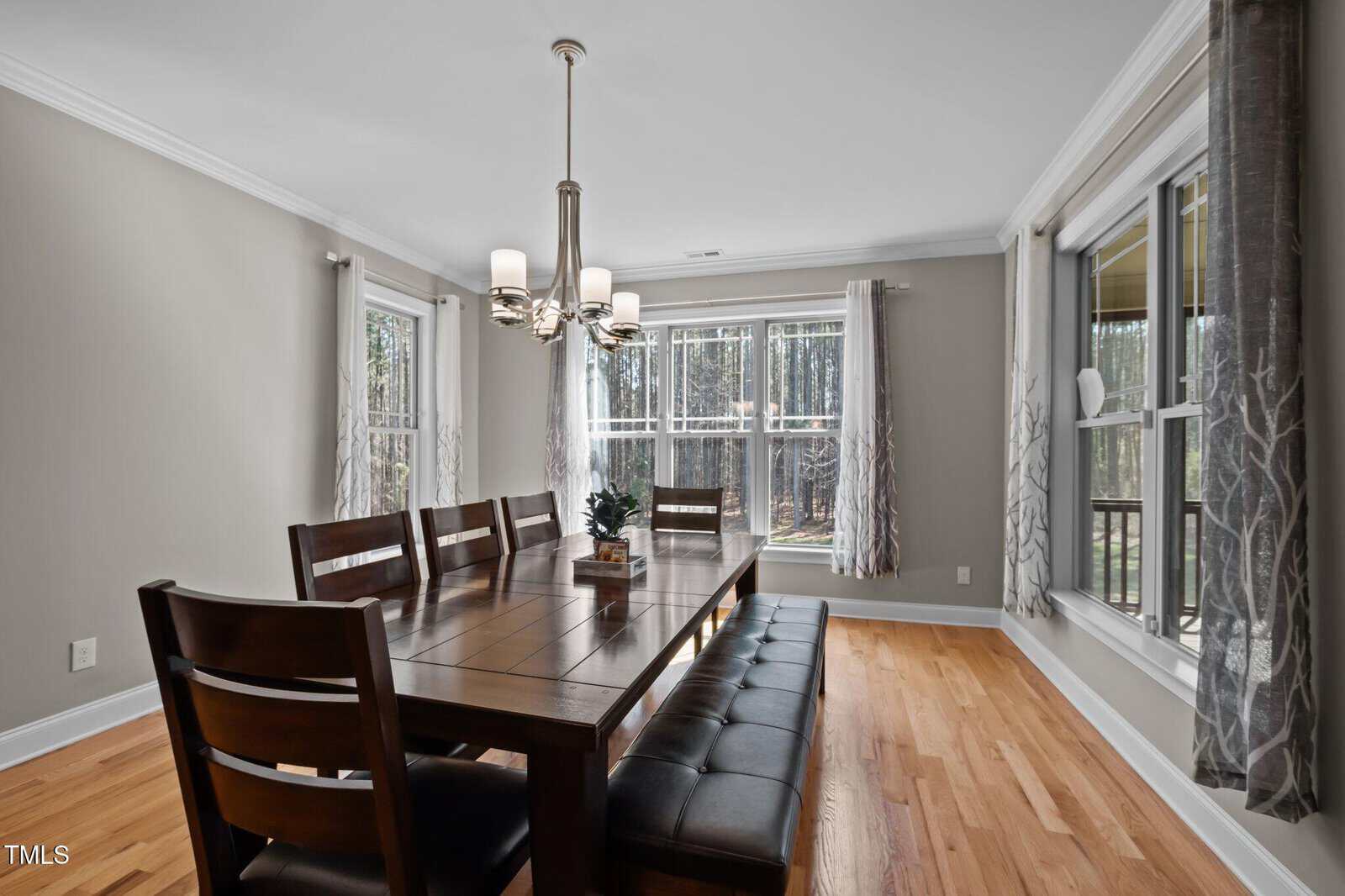 A dining room with a wooden table, six chairs, a cushioned bench, a modern chandelier, hardwood floors, light gray walls, and large windows with curtains letting in natural light.