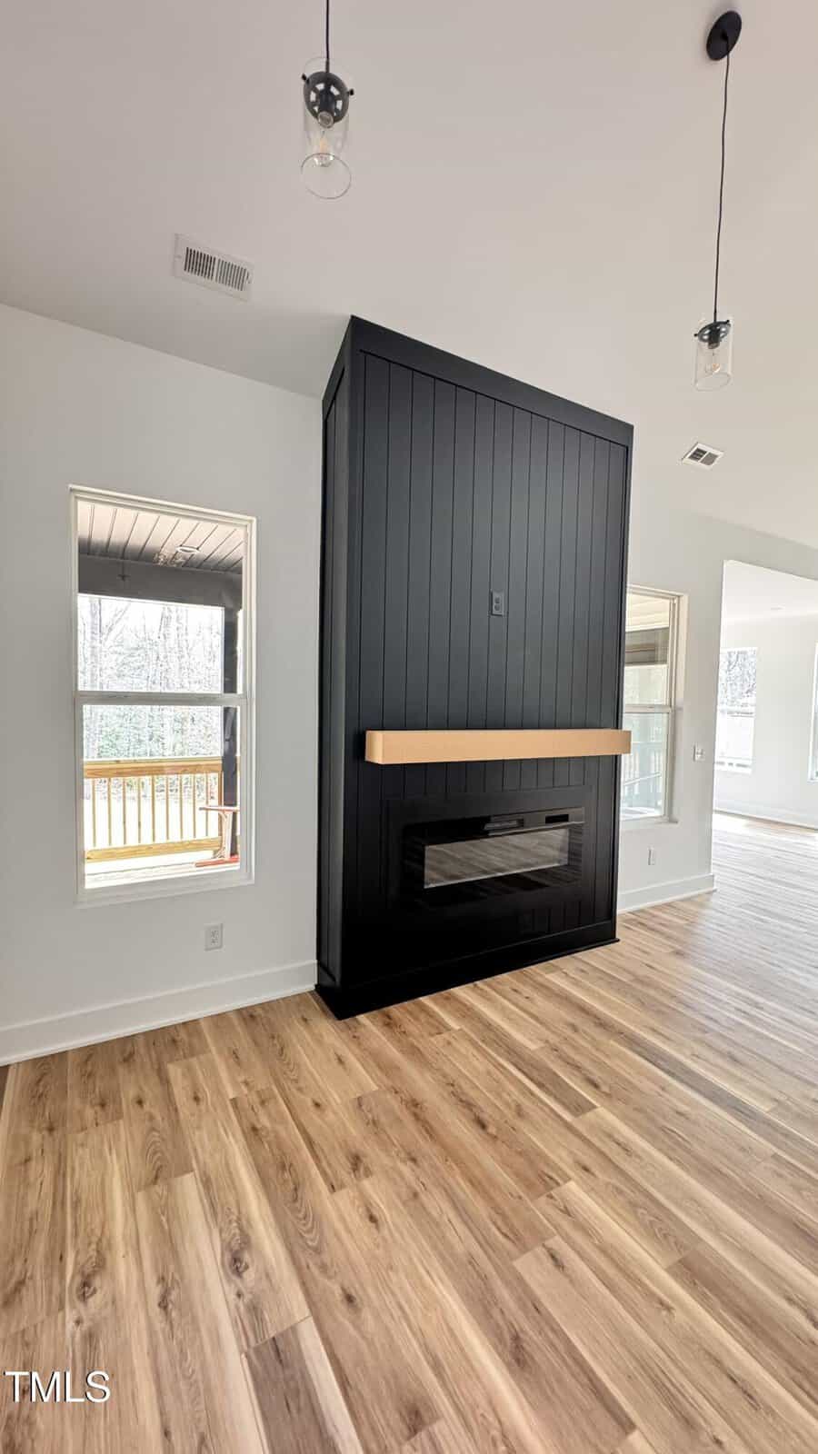 A black shiplap fireplace with a wood mantel is centered in a bright room with light wood floors, large windows, and pendant lights. A covered porch is visible through the window on the left.