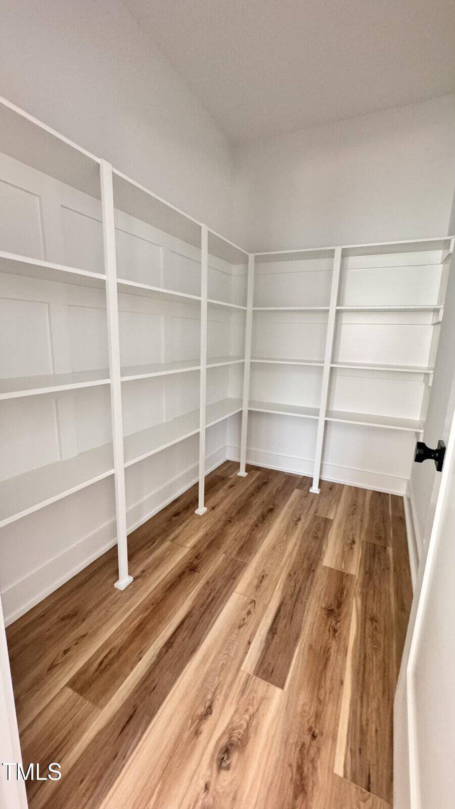 A walk-in pantry with white built-in shelves on three walls and light wood laminate flooring. The walls are white, and the space is empty.