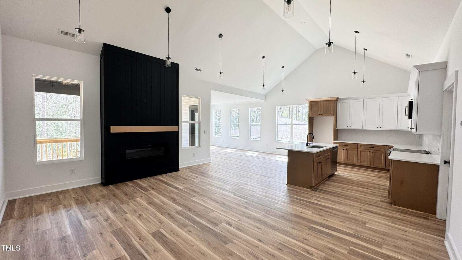 Open-concept living area with vaulted ceiling, black accent wall with fireplace, large windows, wood flooring, and a kitchen with white cabinets, wood island, and pendant lights.