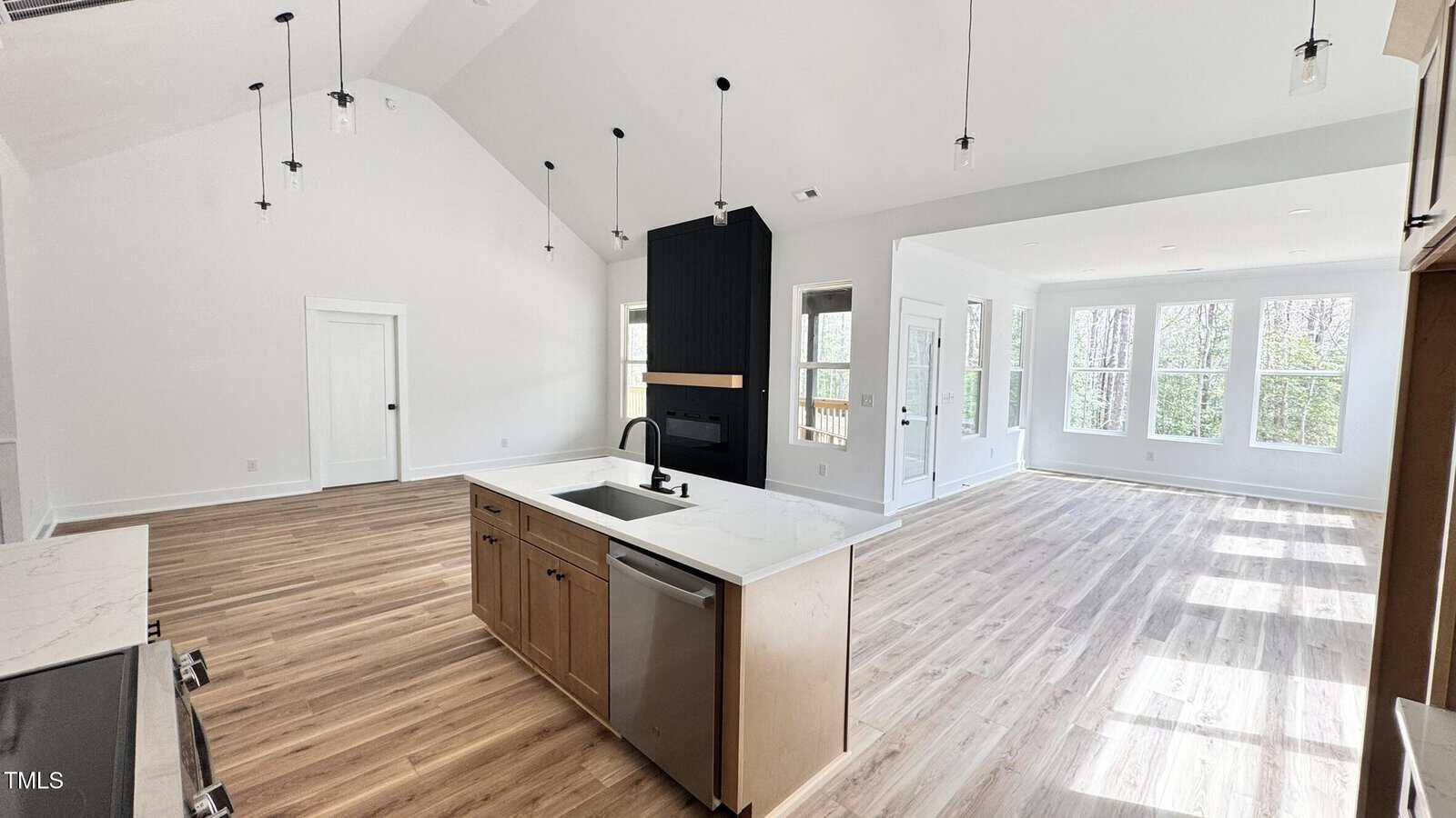 A modern open-concept kitchen and living area with light wood floors, white walls, a central island with a sink and dishwasher, a black accent wall with a fireplace, and large windows letting in natural light.
