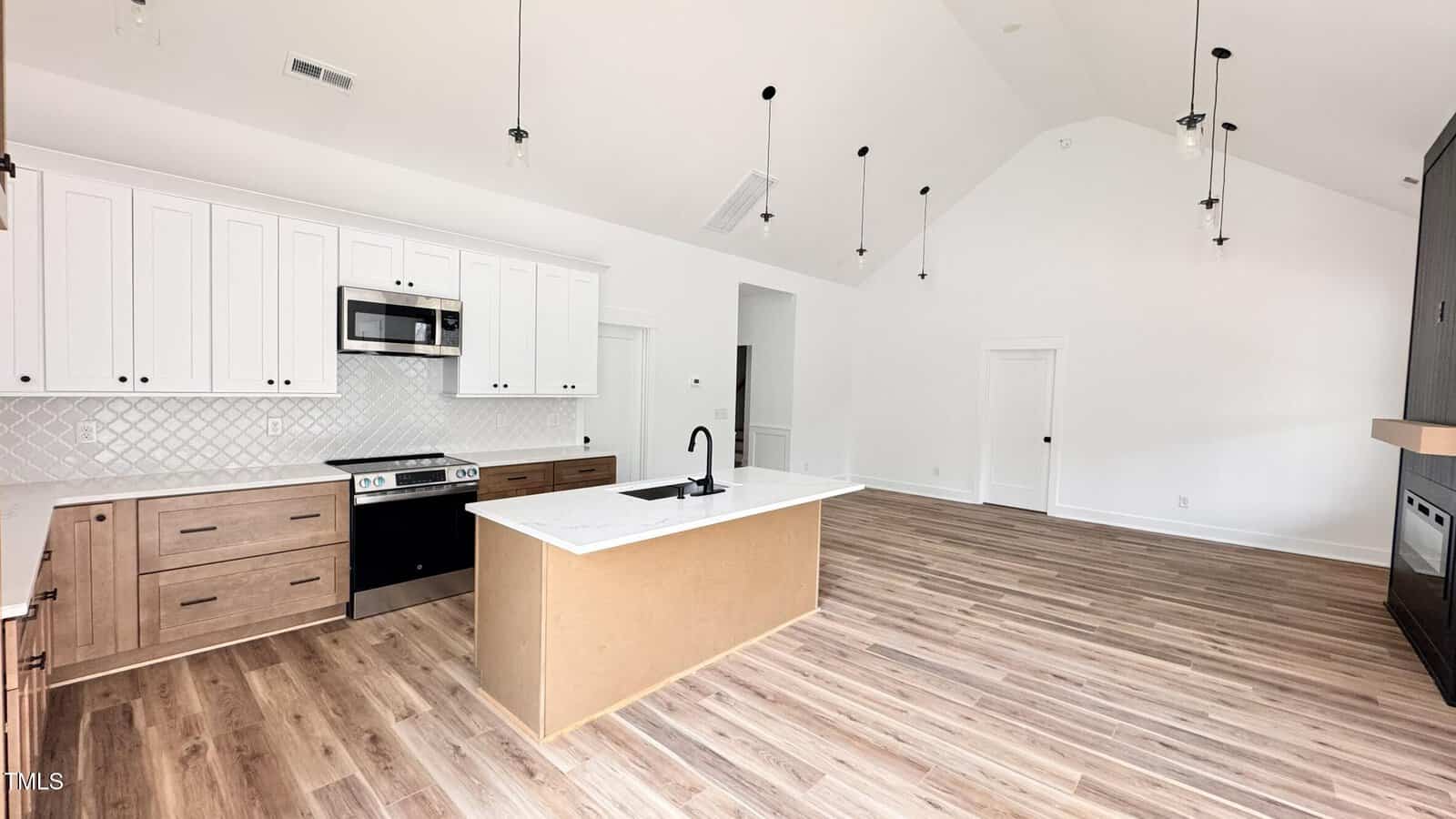 A modern kitchen with white upper cabinets, wood lower cabinets, a center island with a black faucet, stainless steel appliances, and pendant lights, set in an open space with wood flooring and vaulted ceilings.