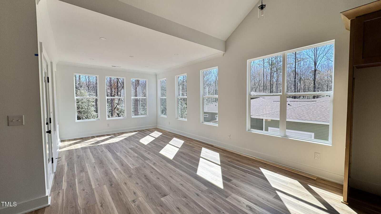 A bright, empty room with large windows on two walls, light wood flooring, and white walls. Sunlight streams in, casting shadows on the floor. Trees and a house roof are visible through the windows.
