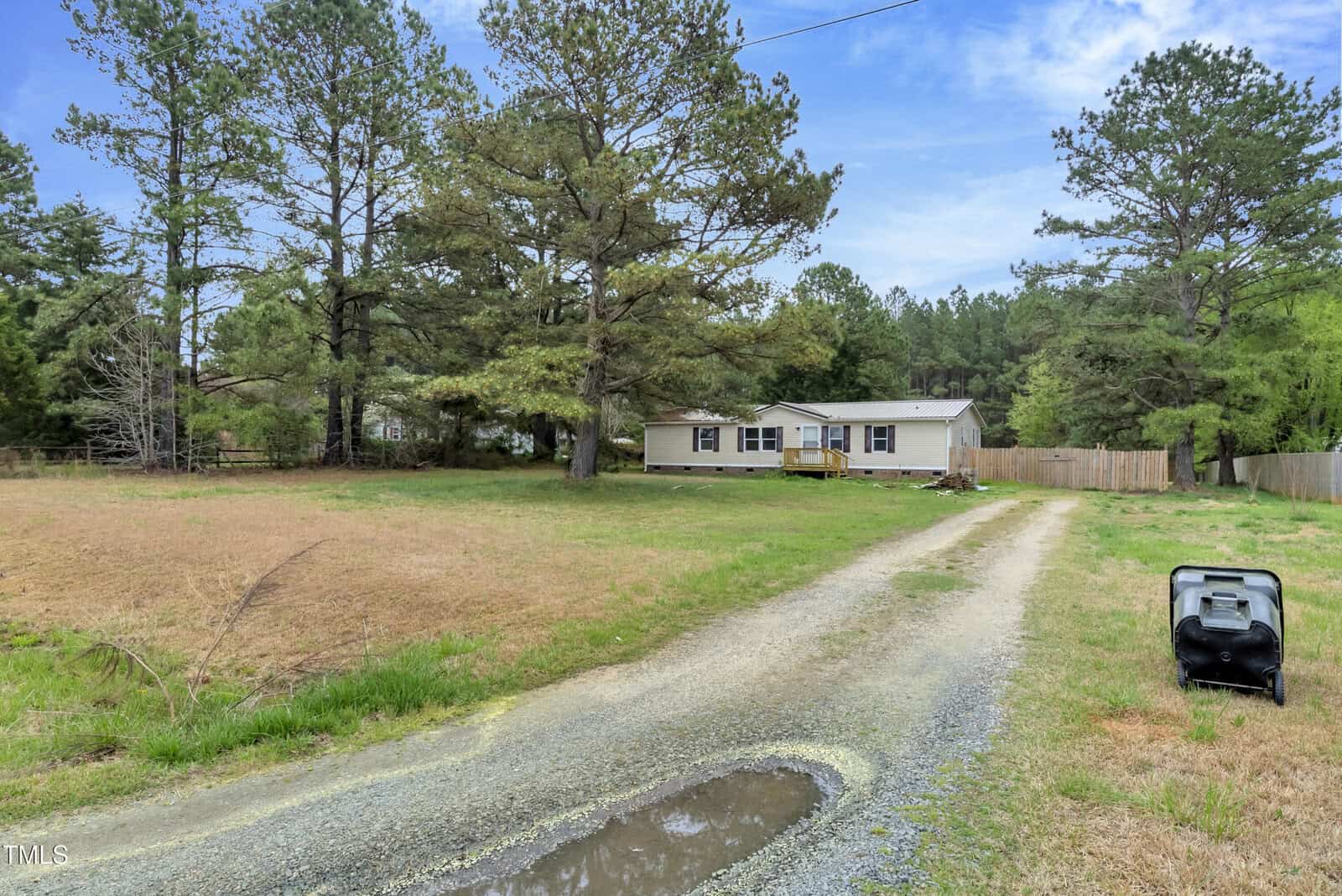 A mobile home on 4296 Tommie Sneed Road in rural Oxford features a gravel driveway. Tall trees and an open grassy field surround it, while a lawnmower rests near the road. The sky above is partly cloudy.