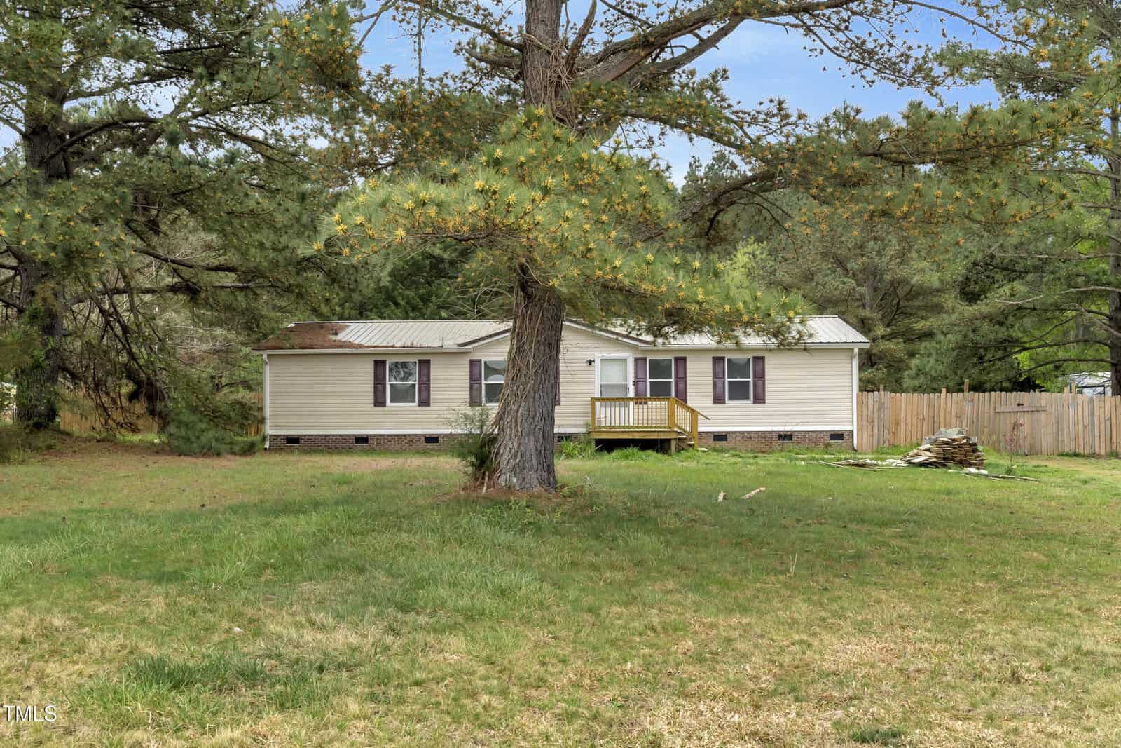 A single-story beige house with a wooden porch sits in a grassy yard. A large tree with overhanging branches is in the foreground. The house is surrounded by a wooden fence and there are trees in the background.