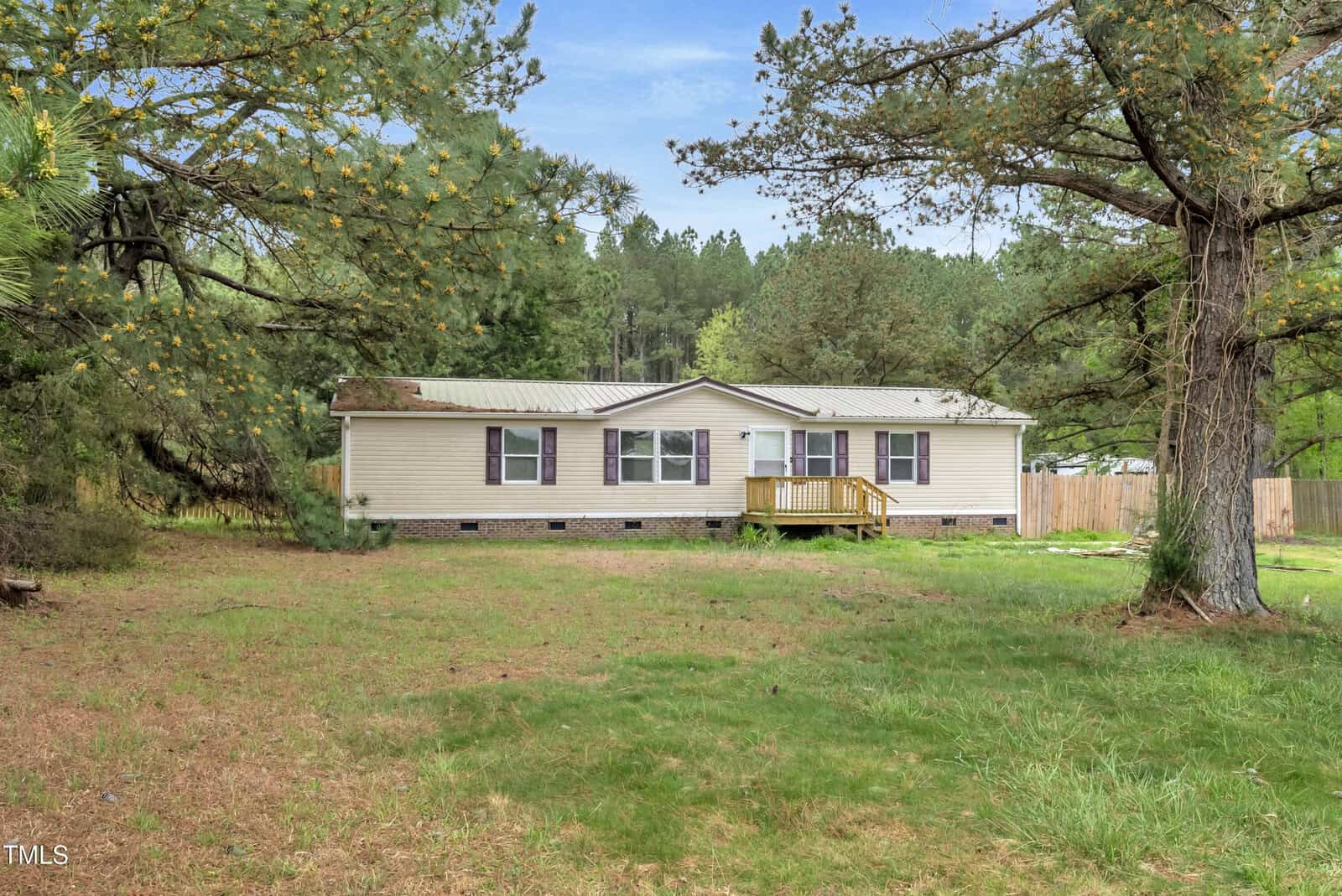 A single-story beige house with a small wooden front porch is surrounded by a grassy yard and tall pine trees. A wooden fence is visible in the background under a clear blue sky.