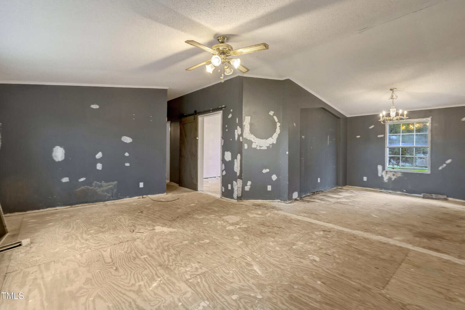 A room with unfinished wood flooring and dark walls, displaying multiple patched areas. A ceiling fan and a light fixture are visible. A window is present on the right wall, allowing natural light. A doorway leads to another room.