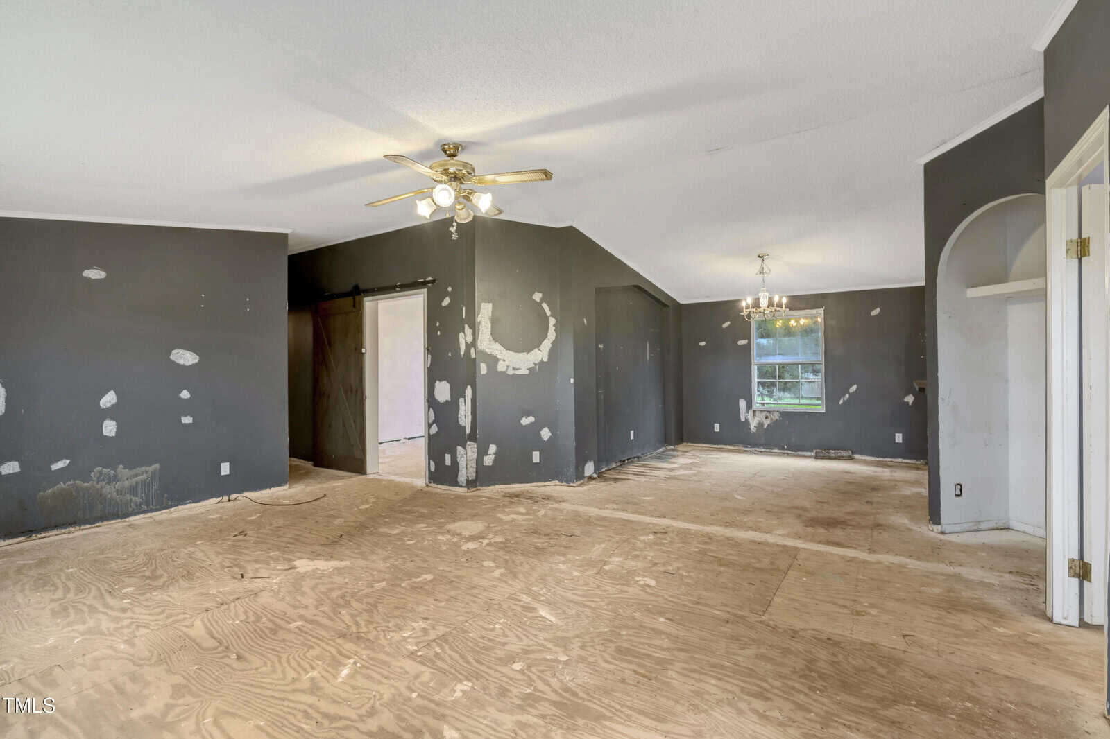 An empty room with unfinished plywood flooring and dark walls with visible patches. Ceiling fans with lights are on, and a window allows natural light in. A doorway leads to another room, and there is a chandelier hanging near the window.