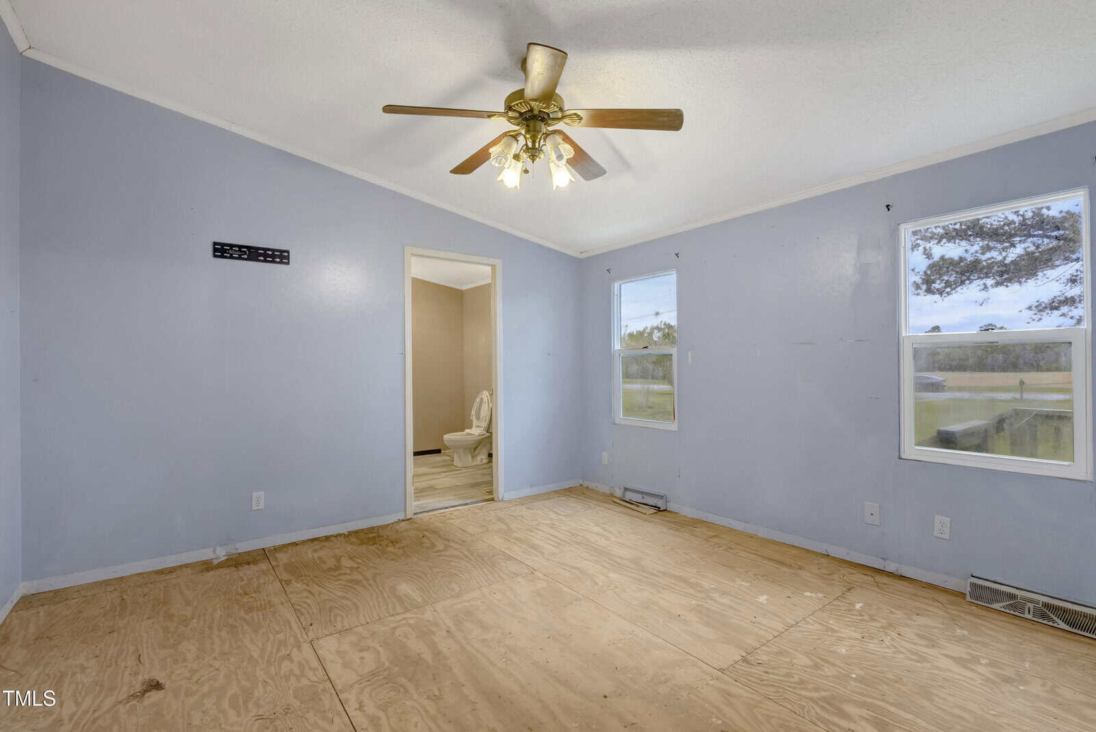 An empty room with light blue walls and a plywood floor. There is a ceiling fan with lights, two windows, and an open doorway leading to a small room with a visible toilet. A TV mounting bracket is on one wall.