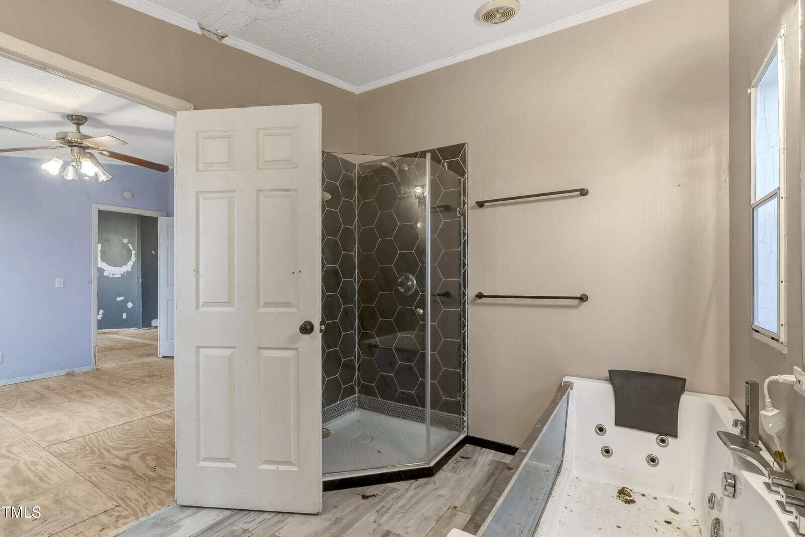 A bathroom with a corner shower enclosed by glass, featuring hexagonal tiles. There is a bathtub with a headrest on the right. The room has beige walls, a door partially ajar, and a visible part of an adjoining room with a ceiling fan.