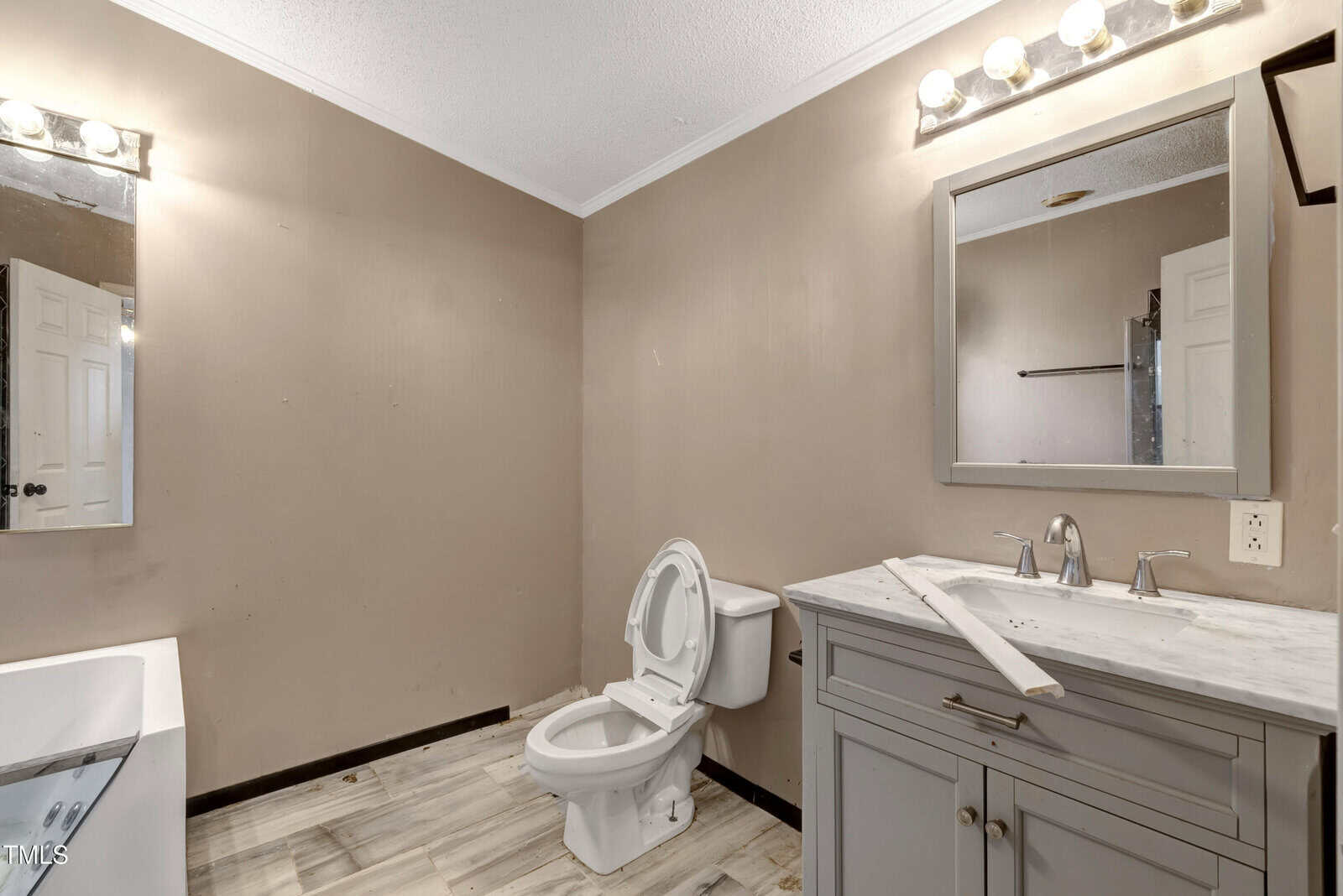 A bathroom with beige walls featuring a toilet, a vanity with a marble countertop, and a mirror. The floor is tiled, and a bathtub is partially visible on the left. The lighting consists of four bulbs above the mirror.