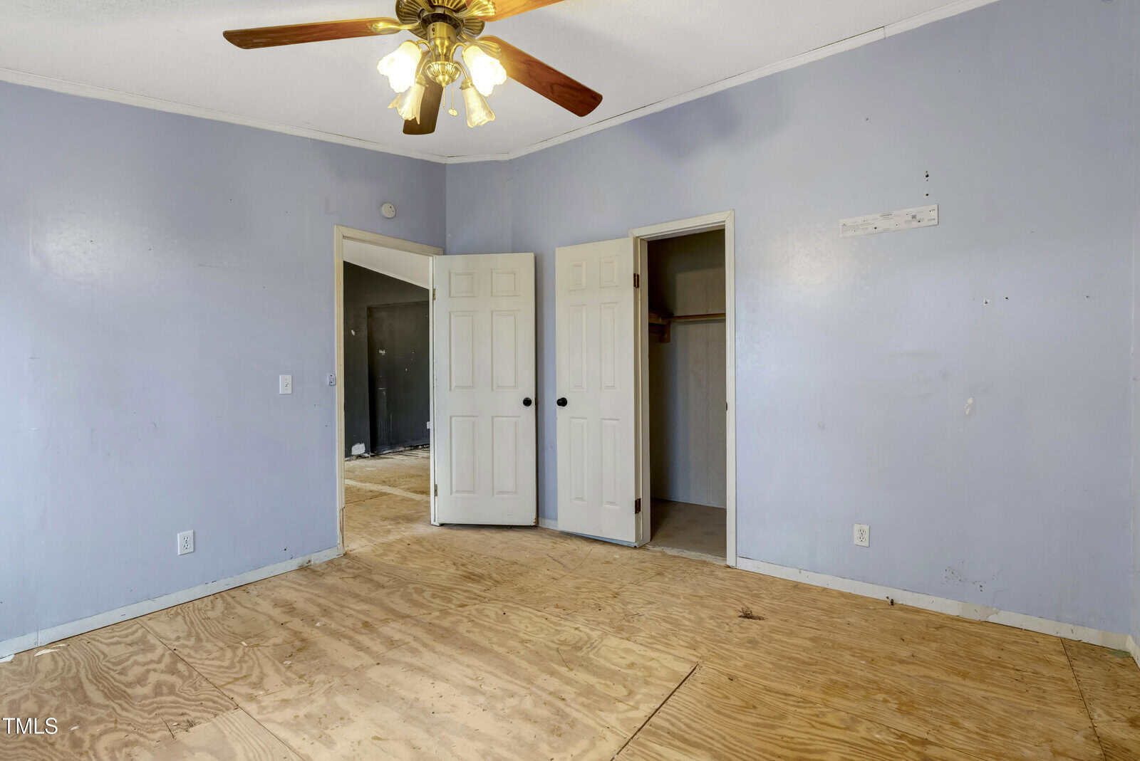 An empty room with light purple walls and a plywood floor. There is a ceiling fan with lights, a white door leading to another room, and a closet with a door slightly open. Some walls show signs of wear.